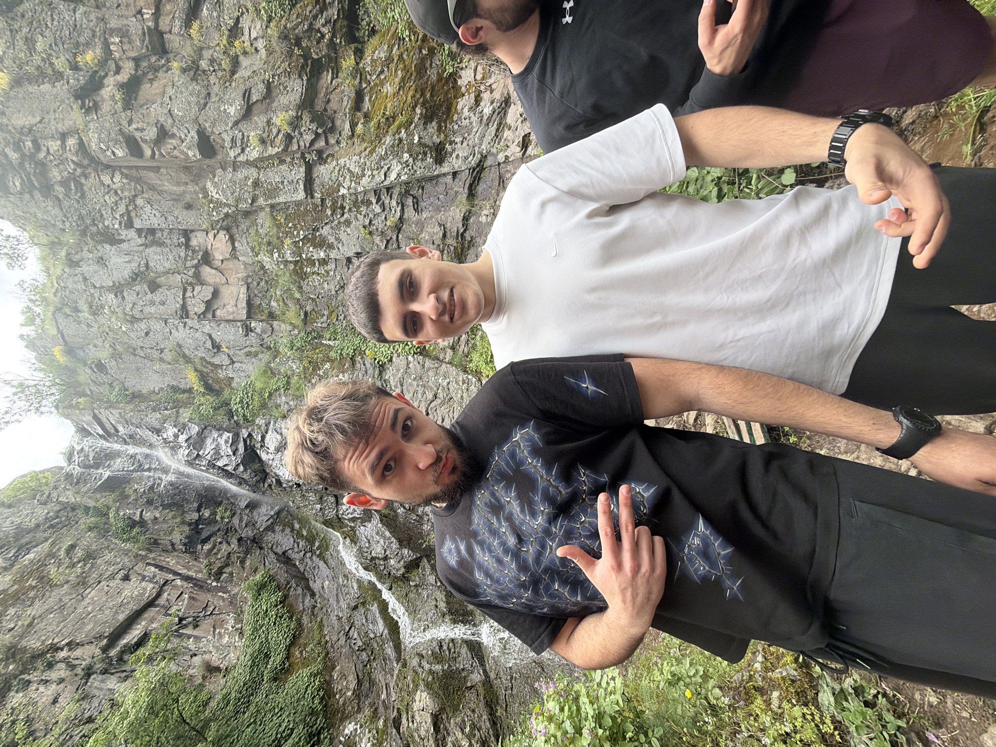 Three men stand in front of a rocky cliff face with a thin waterfall. The man in the foreground wears a black t-shirt with a blue design and black pants, making a hand gesture. Behind him, a second man wears a white t-shirt and black pants. Partially visible behind them is a third man in a black shirt with a black cap. The setting appears to be a natural environment with green vegetation growing on the cliff face. This scene is at the Ustina Waterfall in Bulgaria.