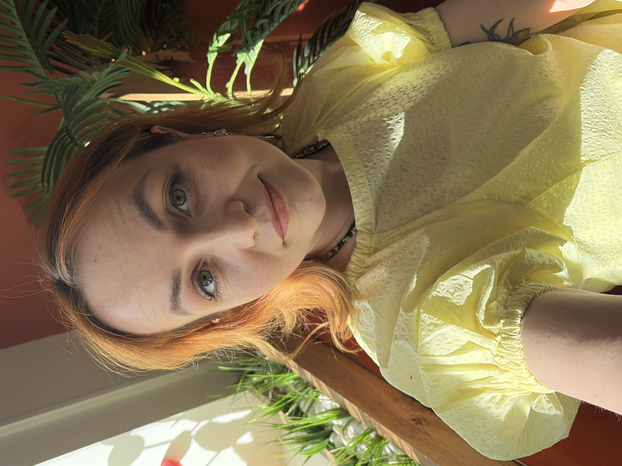 A fair-skinned woman with light auburn hair and green eyes is the primary subject of the image. She is wearing a textured yellow blouse with puffed sleeves. A visible tattoo adorns her arm. A beaded necklace circles her neck. She appears to be positioned near a window, as sunlight floods the scene. Various plants, some with long green leaves, frame the image. The presence of an orange wall suggests an interior space. While the precise location is not explicitly evident from the image, the mention of the city of Chaikovsky, Russia, may indicate that the picture was taken at this location.