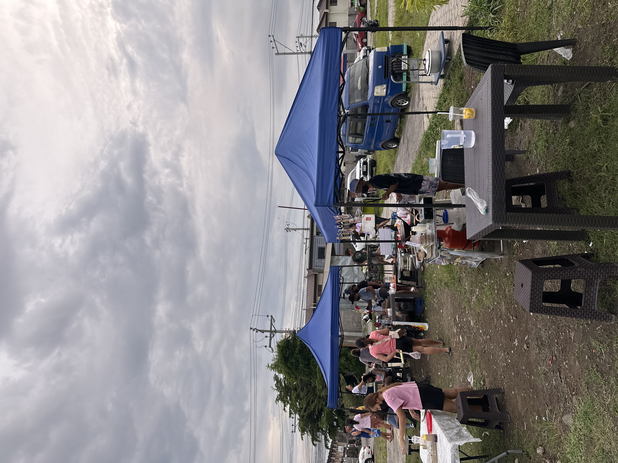 An outdoor market scene in Davao City, Philippines, is captured under a densely overcast, cloudy sky. Several blue pop-up canopies provide shade for multiple temporary stalls set up along a strip of dirt and grass next to what appears to be a paved path or road.
A variety of people, including vendors and customers, are present, mostly standing around the stalls. They are casually dressed in t-shirts and shorts, engaging in various market activities, such as preparing food or browsing items. In the foreground, several dark brown, wicker-patterned plastic tables and stools are visible. One table holds a clear pitcher of water and another pitcher containing a drink with lemon slices.
Further back, under the canopies, other stalls are active with people and assorted items, suggesting food preparation and sales. A small blue Suzuki utility vehicle is parked in the mid-ground, visible through the gaps in the stalls. Its front prominently displays the "SUZUKI" logo and a partial license plate reading "KAM 546". In the background, utility poles with power lines extend across the frame, alongside trees and residential buildings, indicating a semi-urban setting.
The time of day appears to be late morning or afternoon, given the overall brightness despite the heavy cloud cover. The atmosphere is informal and busy, typical of a community market or street food gathering.