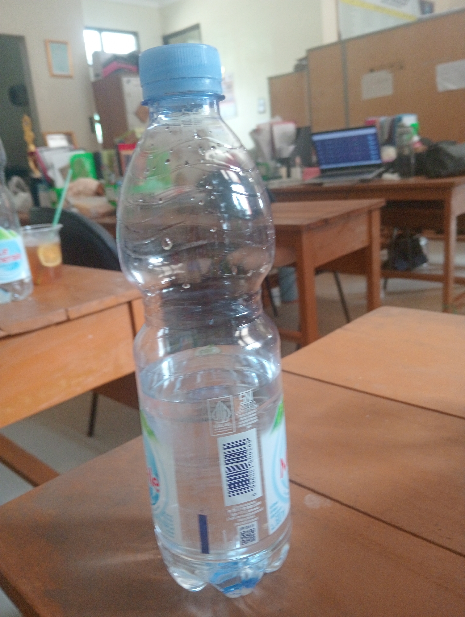 A clear plastic bottle of water with a blue cap stands on a wooden desk. The bottle is partially filled with water, and condensation is visible on its surface. In the background, a laptop is open on another wooden desk, displaying a screen with blue text. Further in the background, office partitions and furniture are visible, suggesting an indoor office or classroom environment. The lighting appears to be natural, coming from windows. The scene is set in a classroom at SMP Negeri 1 Wanasalam, located in Wanasalam, Indonesia.