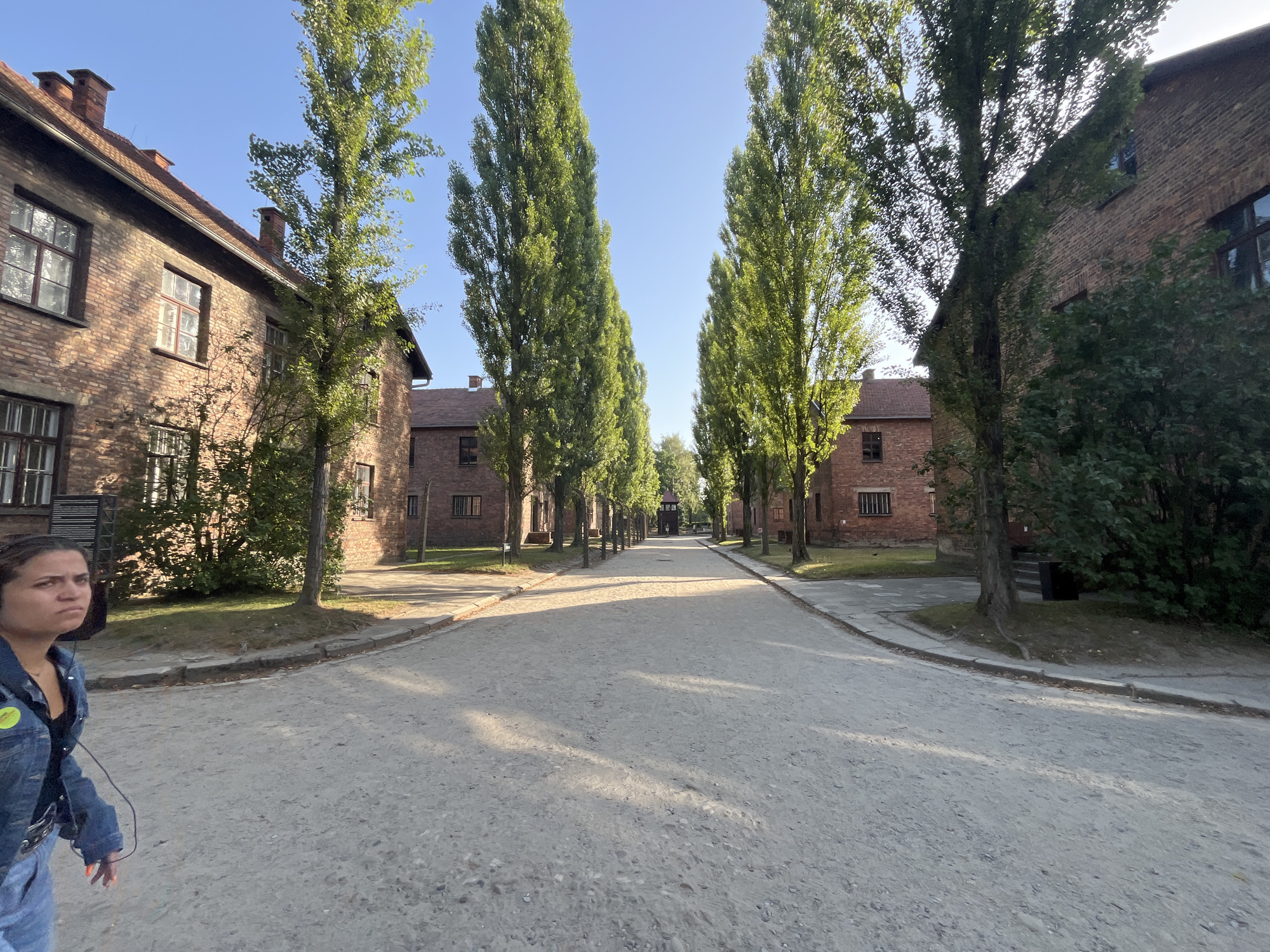 The image captures a path lined with tall trees and brick buildings, characteristic of the Auschwitz-Birkenau State Museum in Oświęcim, Poland. In the foreground, a woman wearing a denim jacket and jeans, with headphones on, is walking towards the right of the frame. Her expression appears somewhat serious. The path is made of gravel and is bathed in sunlight, casting shadows from the trees. The brick buildings on either side have multiple windows and chimneys. The scene conveys a sense of historical significance and solemnity, with the natural elements of trees and sky contrasting with the man-made structures.