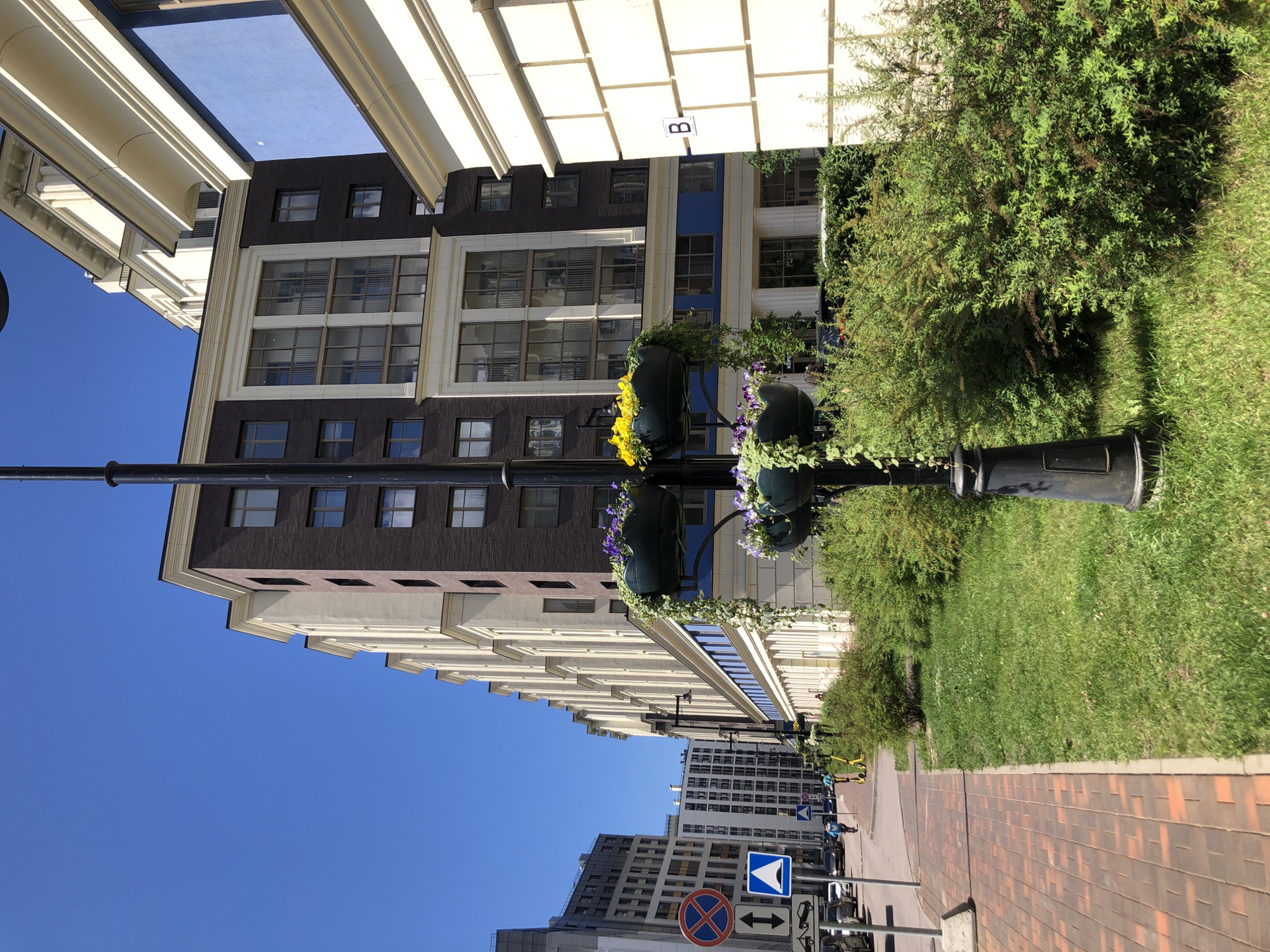 This is a street-level, medium shot of a modern residential complex in St. Petersburg, Russia, on a clear, sunny day. The buildings are contemporary, with dark brick and light-colored facades, featuring numerous windows and balconies. A lamppost with hanging planters filled with purple and yellow flowers stands in the foreground on the right. To the left, a brick sidewalk runs alongside a grassy area with dense green bushes. Road signs indicating no parking and a pedestrian crossing are visible on the left side of the street. Cars are parked along the road, and a few people are seen cycling and riding electric scooters. The architectural style suggests a new development in an urban environment.
