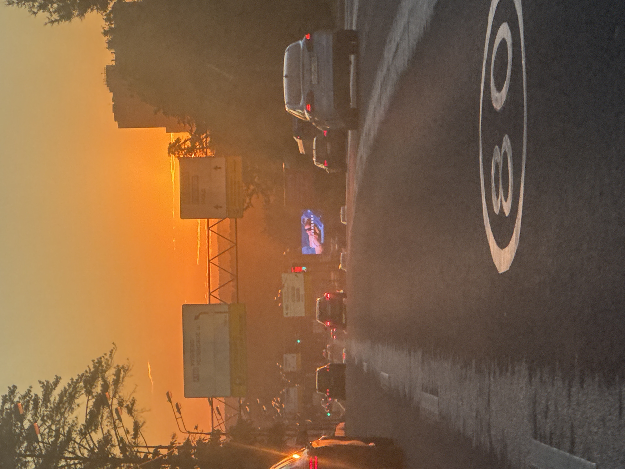 The image depicts a street scene in Moscow, Russia, during sunset or sunrise, indicated by the warm, orange glow of the sky. The perspective is from a moving vehicle, looking towards the right side of the road. The road is wet, suggesting recent rain or high humidity, and there are several cars on the road, some with their brake lights illuminated. A prominent speed limit sign with the number "80" inside a white circle is visible on the asphalt.
On the left side of the image, there are trees with silhouetted branches against the bright sky, and some buildings are visible in the distance, partially obscured by atmospheric haze. Several road signs are also present, though the text on them is not clearly legible. One sign appears to be a billboard with an image of a person. The overall atmosphere is hazy and somewhat dreamlike due to the lighting conditions and the motion blur from the camera. The scene conveys a sense of travel and urban movement during twilight hours.