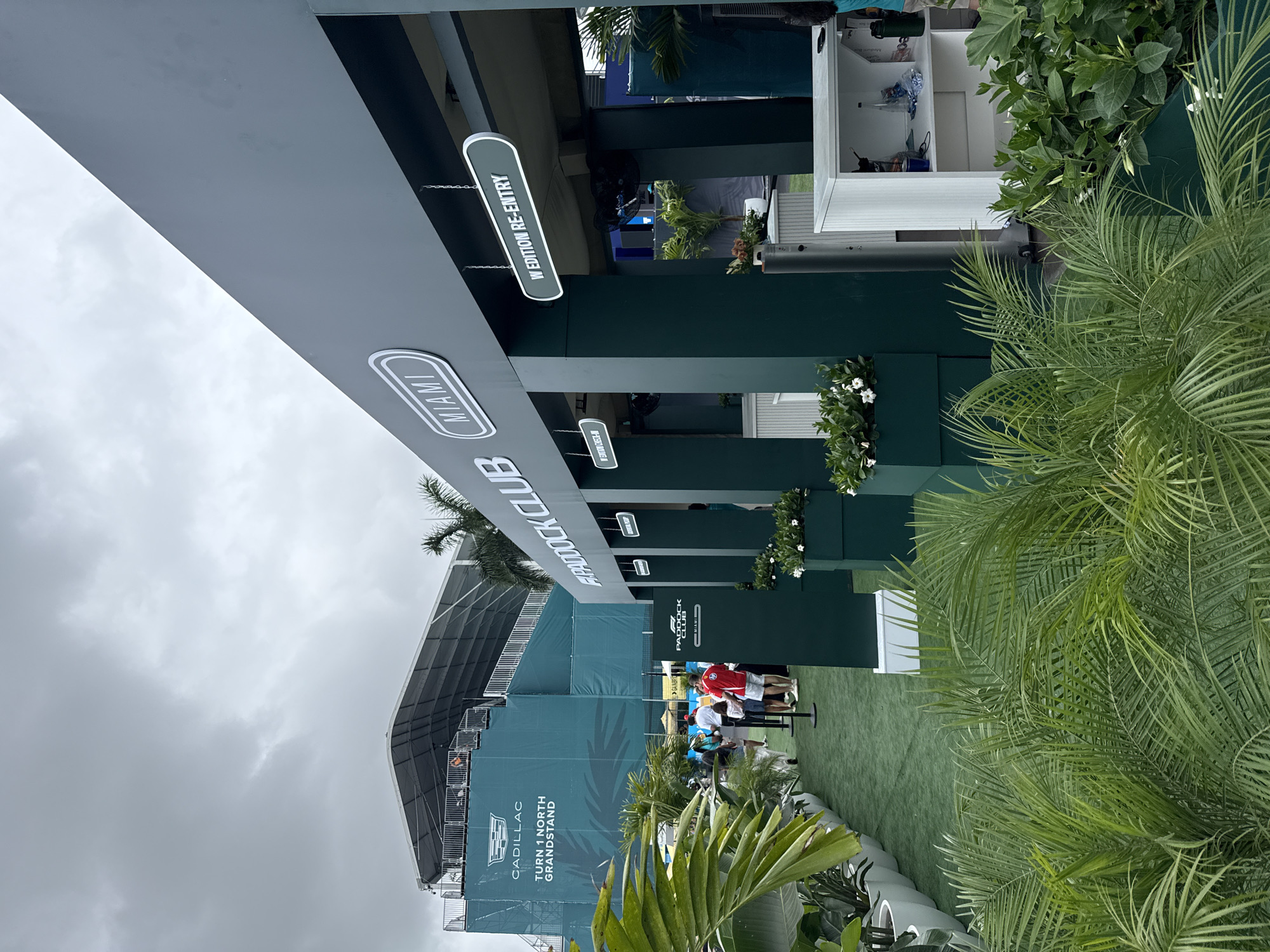 The image shows the Paddock Club entrance at the Miami International Autodrome in Miami Gardens, Florida. The sky is overcast. A large grey awning with the "Paddock Club" logo hangs over the entrance. Signs indicate "Miami" and "W Edition Re-Entry." People are seen queuing at the entrance. A large structure for the Cadillac Turn 1 North Grandstand is in the background. Lush greenery including palms is in the foreground.