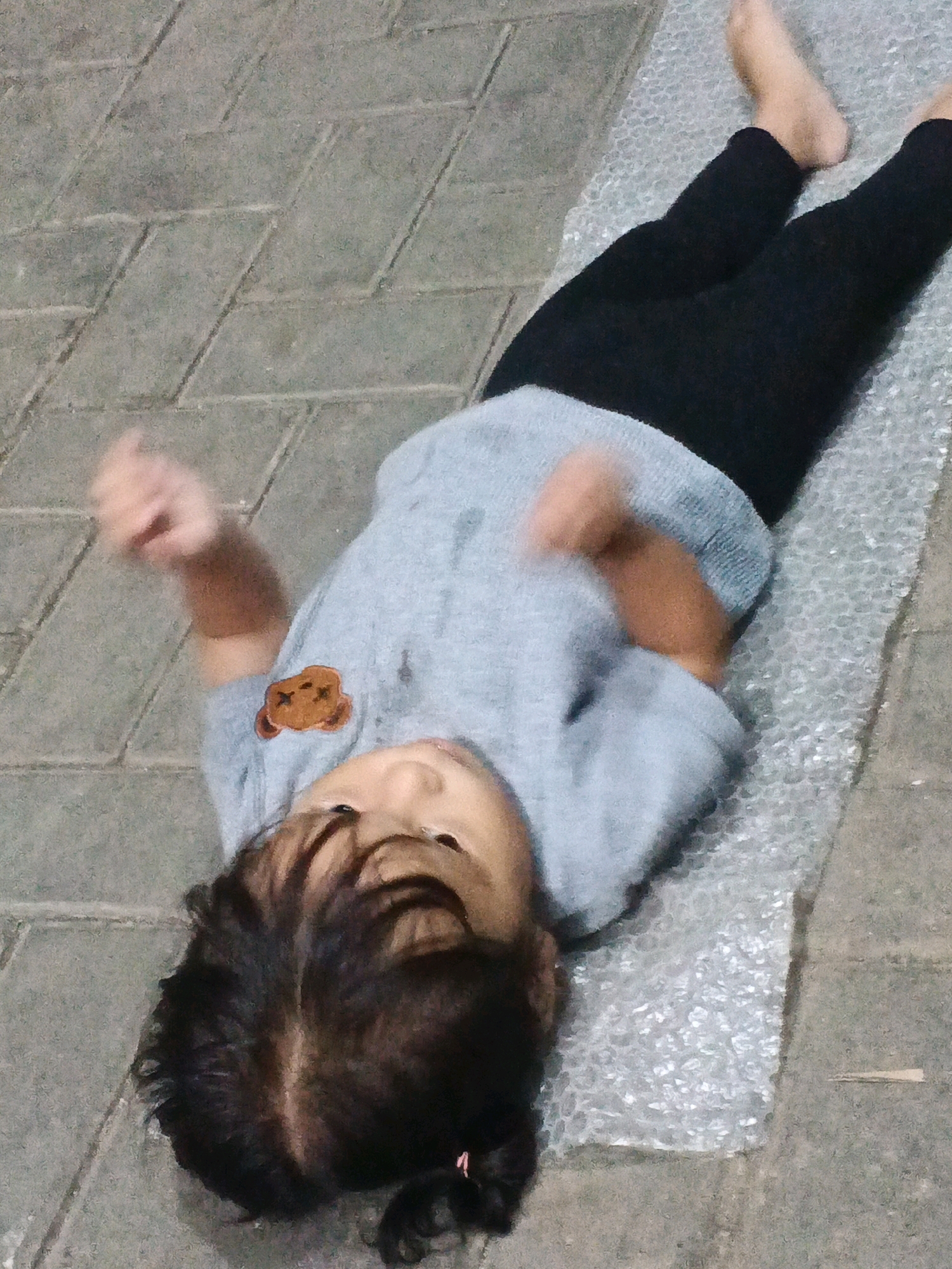 A young child is lying on their back on a textured surface, likely a padded mat or bubble wrap, which is placed on a paved stone ground. The child is wearing a light blue long-sleeved shirt with a brown bear applique on the left chest and black leggings. Their head is turned to the left, and their face is partially visible, with dark hair styled in a way that suggests it might be tied back. One arm is raised and bent, appearing to be in motion. The background shows the greyish, textured paving stones of an outdoor area. The scene is lit, suggesting it might be daytime or under artificial light. The location is identified as Kadu, Indonesia, implying an outdoor setting within that city.
