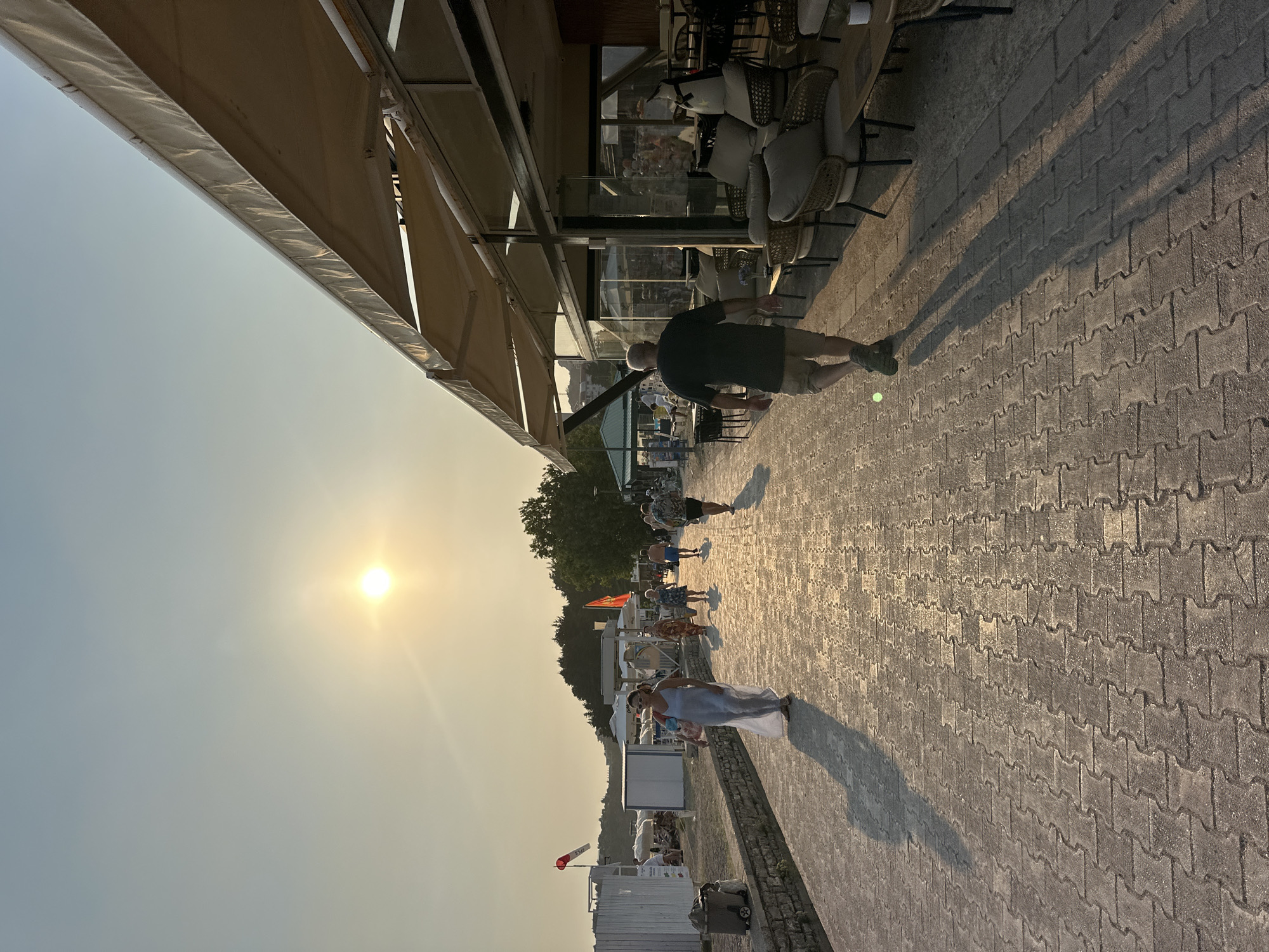 This is an outdoor, eye-level, full shot of a paved walkway along a beach at dusk. The sun is setting and casting long shadows. On the right side of the image, a restaurant with outdoor seating is visible. Several people are walking along the path. On the left side of the path, beach umbrellas and chairs are set up. The location appears to be the promenade in Boreti, Montenegro.