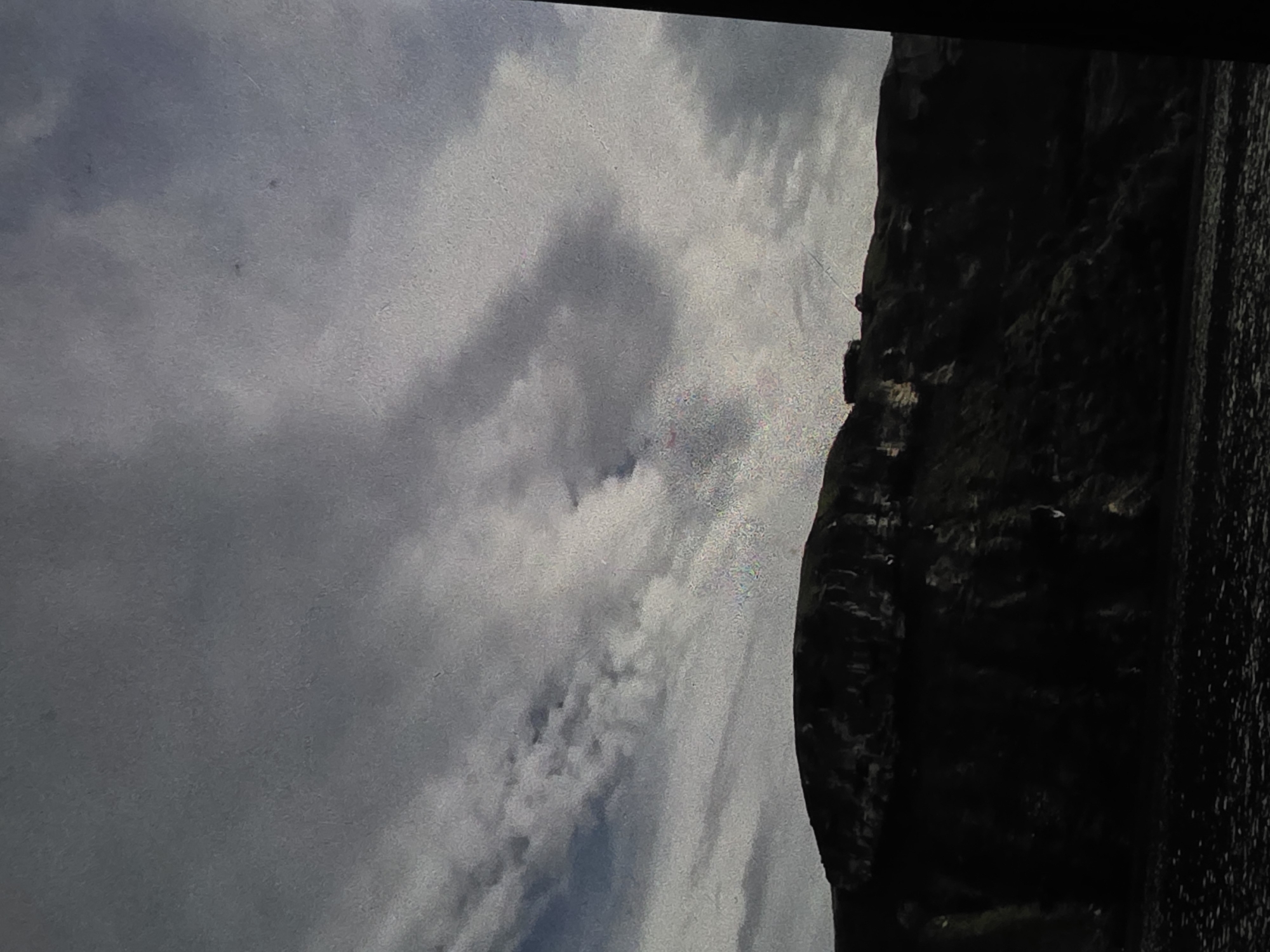 The image depicts a dark, mountainous landscape under a cloudy sky. The foreground is a dark, rocky terrain, possibly a beach or a scree slope. Rising behind it is a steep, dark mountain or cliff face, casting deep shadows. The summit of the mountain is silhouetted against the sky and appears to have some structures on it, possibly buildings or ruins. The sky is filled with dramatic, heavy clouds, with patches of lighter sky visible through them. There are no people or animals visible in the image. The overall impression is one of a dramatic and somewhat bleak natural landscape.
Regarding the location in the city of Dubno, Ukraine, there is no visual information in the image that can definitively place it within Dubno or any specific location. The image shows a generic mountainous or hilly coastal environment.