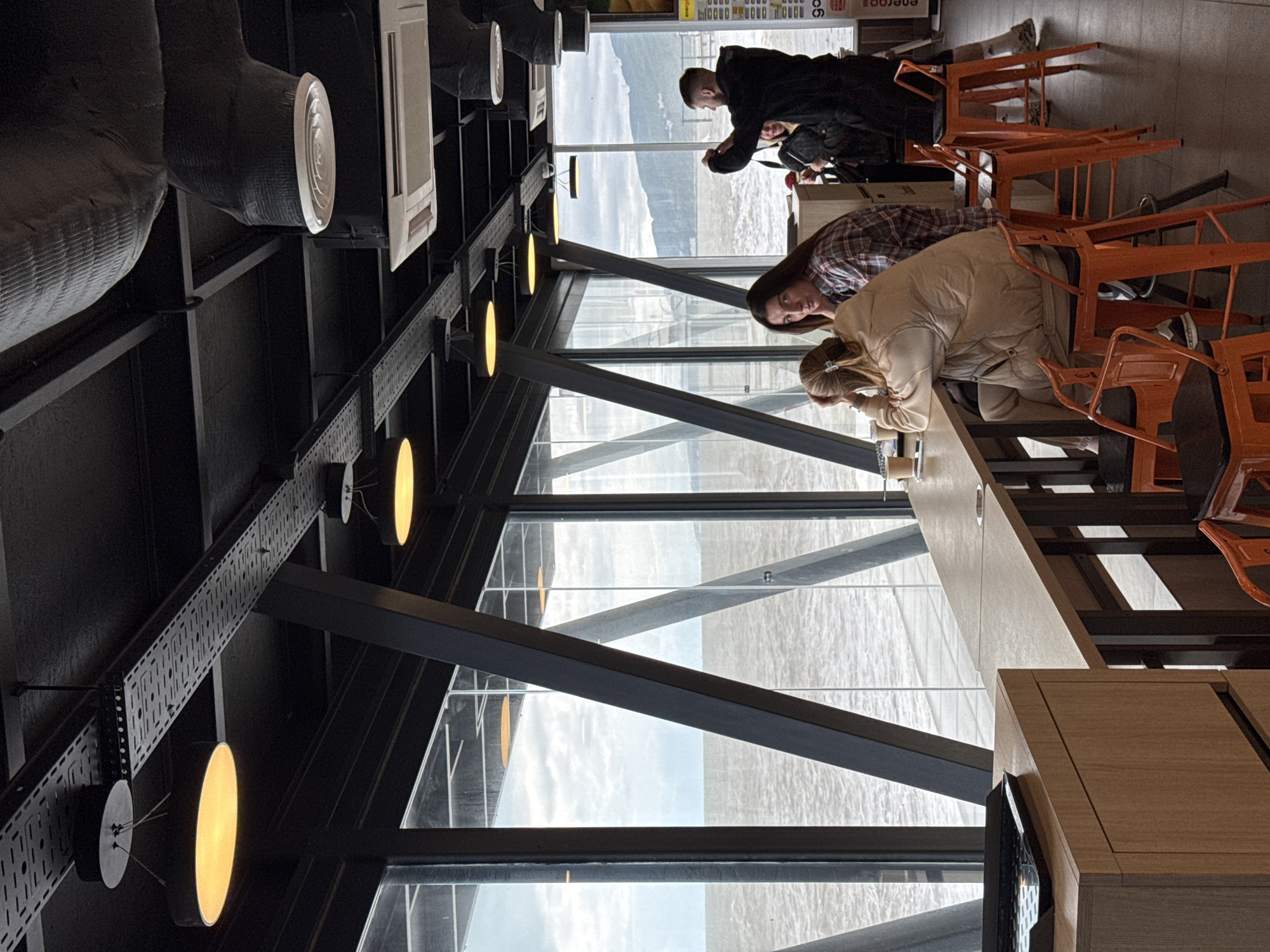 People are sitting at a counter in a cafe or restaurant. The location has large windows that offer a view of what appears to be a body of water, possibly the Black Sea, indicating that the location might be in Sochi, Russia. Three individuals are visible: one is looking at a phone, while the other two are facing the windows. The interior of the space includes exposed black steel beams and lights. Orange chairs line the counter.