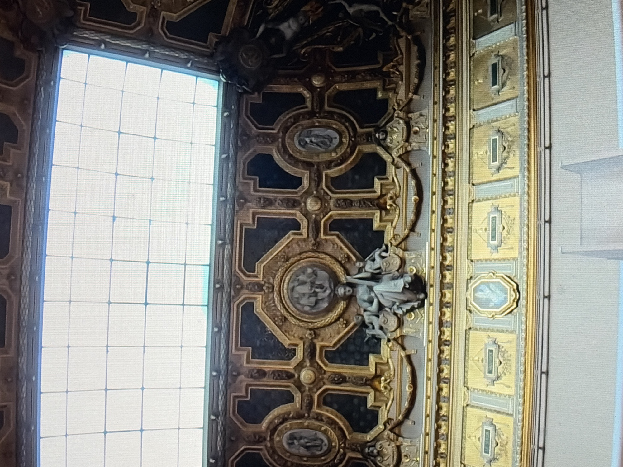 The image shows a detailed view of an ornate interior ceiling. A large grid of square translucent panels forms a skylight at the top, admitting bright light. Below the skylight, the ceiling is elaborately decorated with dark, possibly black, panels framed by intricate gold ornamentation. These gold elements feature geometric shapes, floral motifs, and circular medallions containing bas-relief sculptures.
Prominently displayed in the lower central portion of the decorated ceiling is a bronze-like sculpture of a seated female figure with three children. The woman appears to be holding a scepter. Flanking this central sculpture are smaller oval medallions containing figures and more elaborate gold framing. Beneath this main decorative frieze is a band of alternating rectangular and octagonal panels, also adorned with gold detailing. The rectangular panels contain what appear to be inscriptions or names, with one central octagonal panel featuring a blue and gold crest or emblem. The lower part of the image shows a plain white wall or architectural element. The overall impression is one of opulence and artistic craftsmanship, likely within a public building, museum, or grand hall. The specific location is Unknown, Unknown.