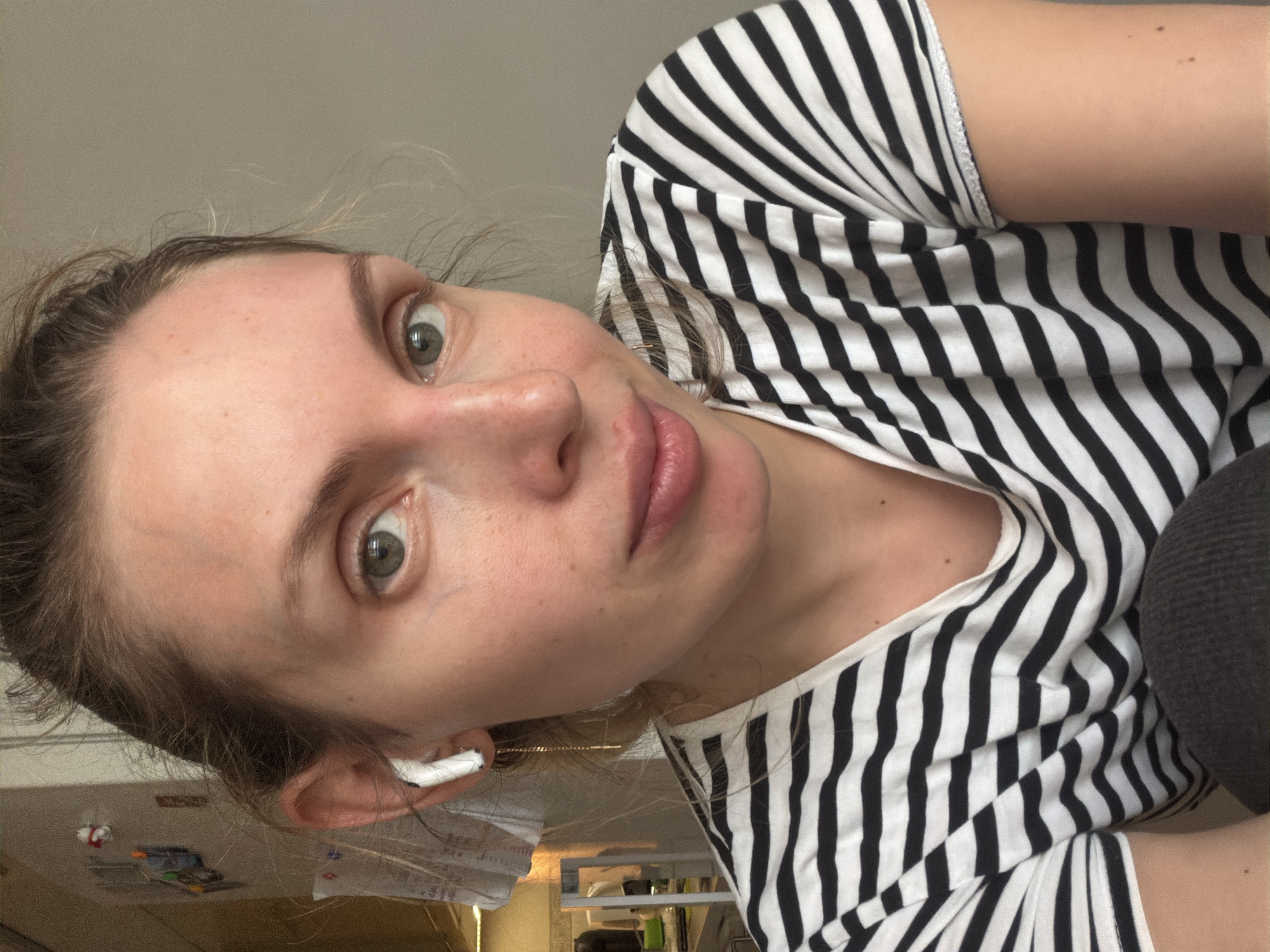 A close-up, indoor selfie of a fair-skinned woman with green eyes. She is wearing a black and white striped shirt and an earbud in her left ear. The background is out of focus, but shows a table with various items, likely in a kitchen. There is no clear indication that the location is Budva, Montenegro. The focus is on the woman's face.