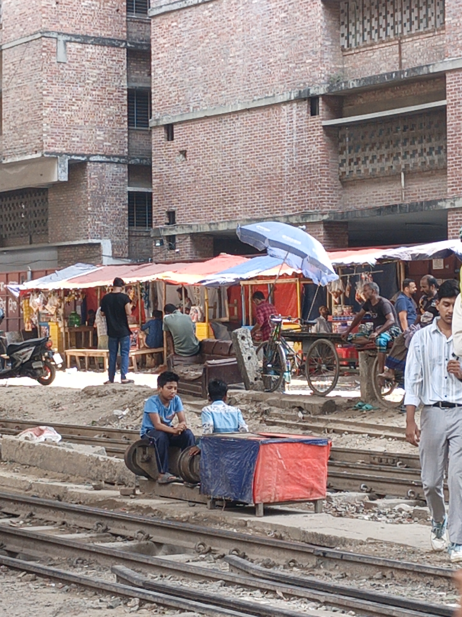 The image depicts a bustling scene in Dhaka, Bangladesh, likely near a railway station. A red brick building serves as the backdrop for a makeshift marketplace under colorful awnings. Numerous individuals are present, including vendors tending to their stalls, customers browsing, and pedestrians passing by. Two young boys are seated on what appears to be railway equipment on the tracks, while another man walks along the tracks. A motorcycle and a rickshaw are visible among the market stalls. The scene is set on a railway track environment.