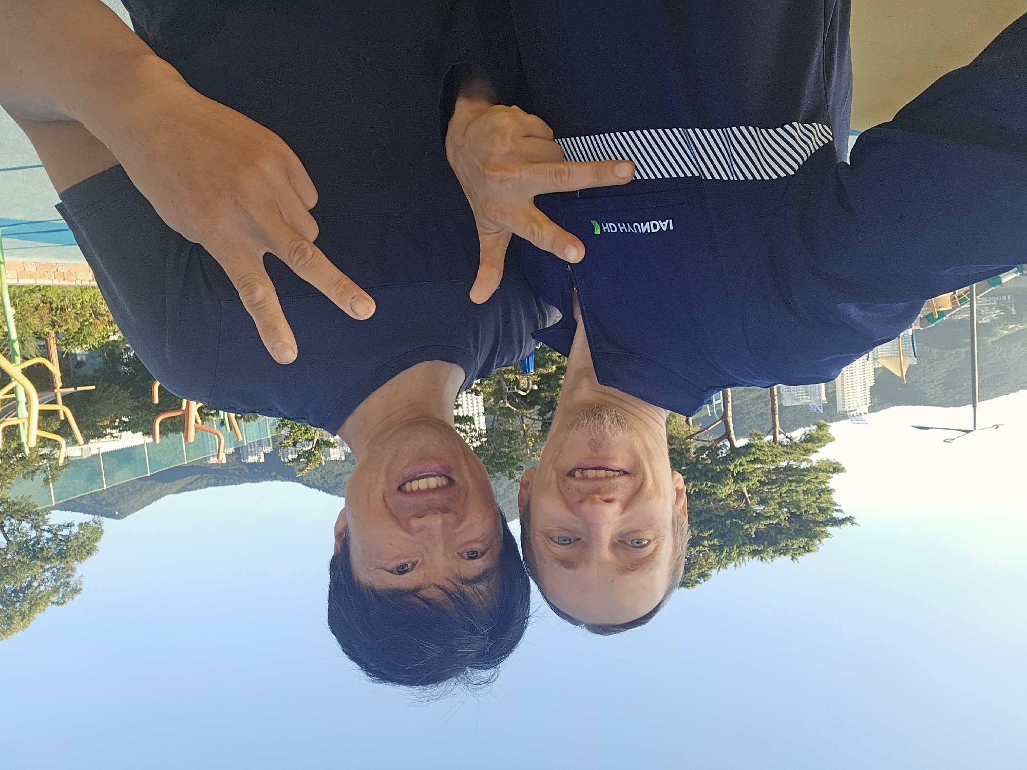 Two men are taking a selfie outdoors in Geoje-si, South Korea. The man on the left is wearing a dark blue long-sleeved shirt with a white striped band across the chest and a logo that appears to be reversed and says "HYUNDAI". He has light skin, blond hair, and a goatee. The man on the right has dark hair and East Asian features. Both men are smiling and making a peace sign with their fingers. They are standing in front of a bright blue sky, some trees, buildings, and mountains in the background. There are also some colorful playground structures visible. The lighting suggests it is daytime, likely morning or afternoon.