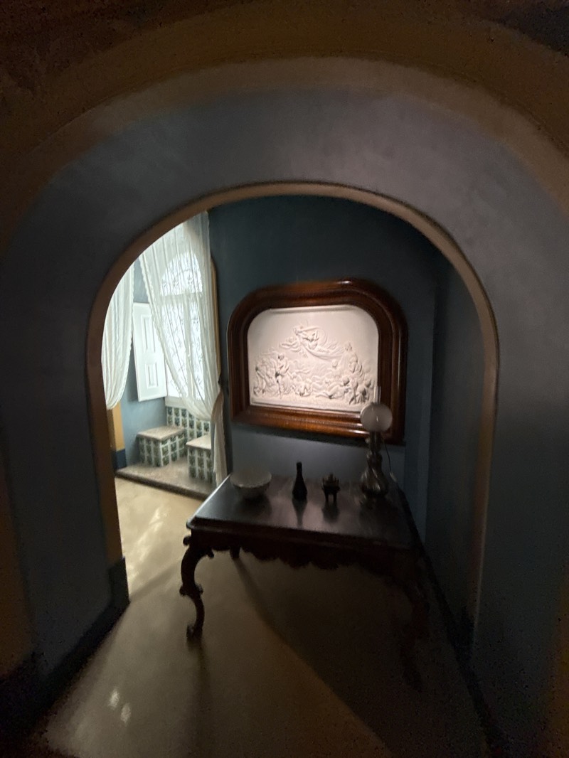 View through an archway of a room in Quinta da Regaleira, Sintra, Portugal. Inside the room is a dark wood table with a white bowl, small dark bottle, and a silver-colored lamp. Above the table is a framed, white bas-relief artwork. The room beyond contains a window with white curtains and two tiled steps.