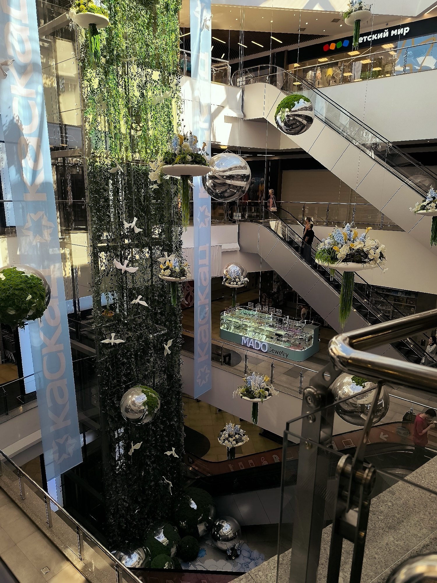 This image captures an interior view of a multi-level shopping mall. In the foreground, a polished metal railing and glass partition suggest a balcony or walkway overlooking the lower levels. Dominating the center of the frame is a tall, cylindrical structure covered in lush green foliage, adorned with white bird-shaped decorations and suspended floral arrangements. Large, reflective spheres are also visible, hanging at various heights and reflecting the surrounding environment.
On the right side of the image, escalators are in motion, with several individuals ascending and descending. One person in a dark top and dark pants is going up, while another in a red shirt is visible on a lower level, looking at their phone.
In the background, store signage can be seen, including "ДЕТСКИЙ МИР" (Children's World) and "MADO Jewelry." The mall appears to be modern, with clean lines and ample lighting. The overall impression is one of a well-maintained and vibrant commercial space. The location is identified as being in the city of Qarağandı, Kazakhstan.