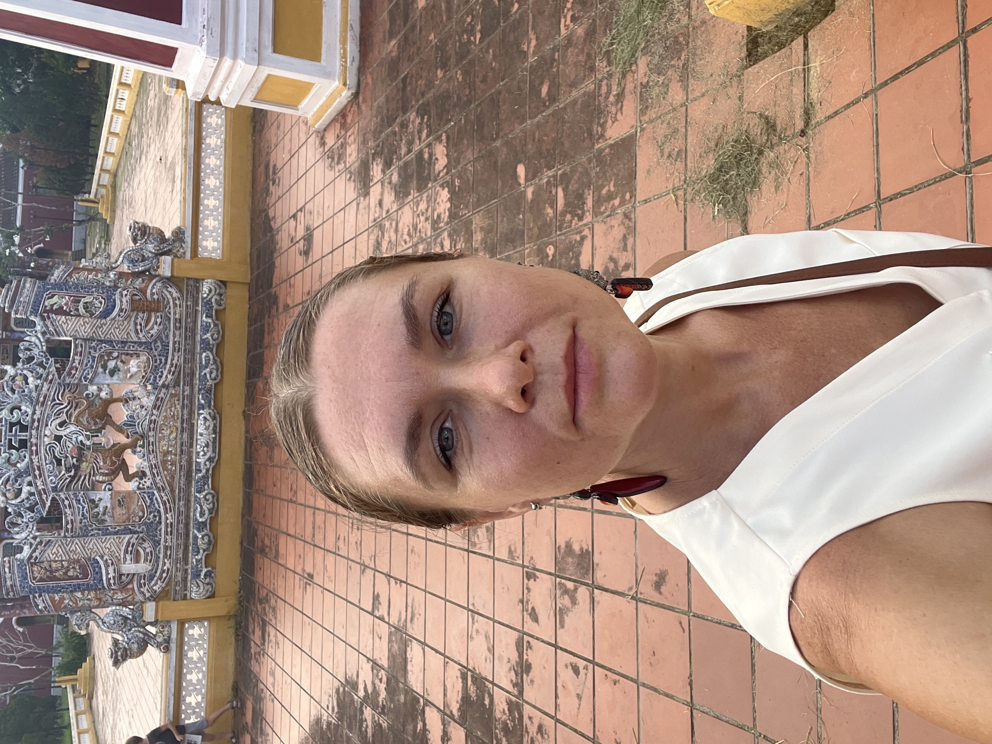 A woman with blonde hair pulled back stands outdoors, taking a selfie. She is wearing a white sleeveless top and large, dark red earrings. Her face is turned towards the camera, and she has blue eyes. Behind her, a courtyard is paved with terracotta tiles. In the background, there is an ornate, colorful tiled wall with dragon motifs, a yellow and white pillar, and some greenery. The scene appears to be within the Imperial City of Hue, a UNESCO World Heritage site, rather than Hoi An.