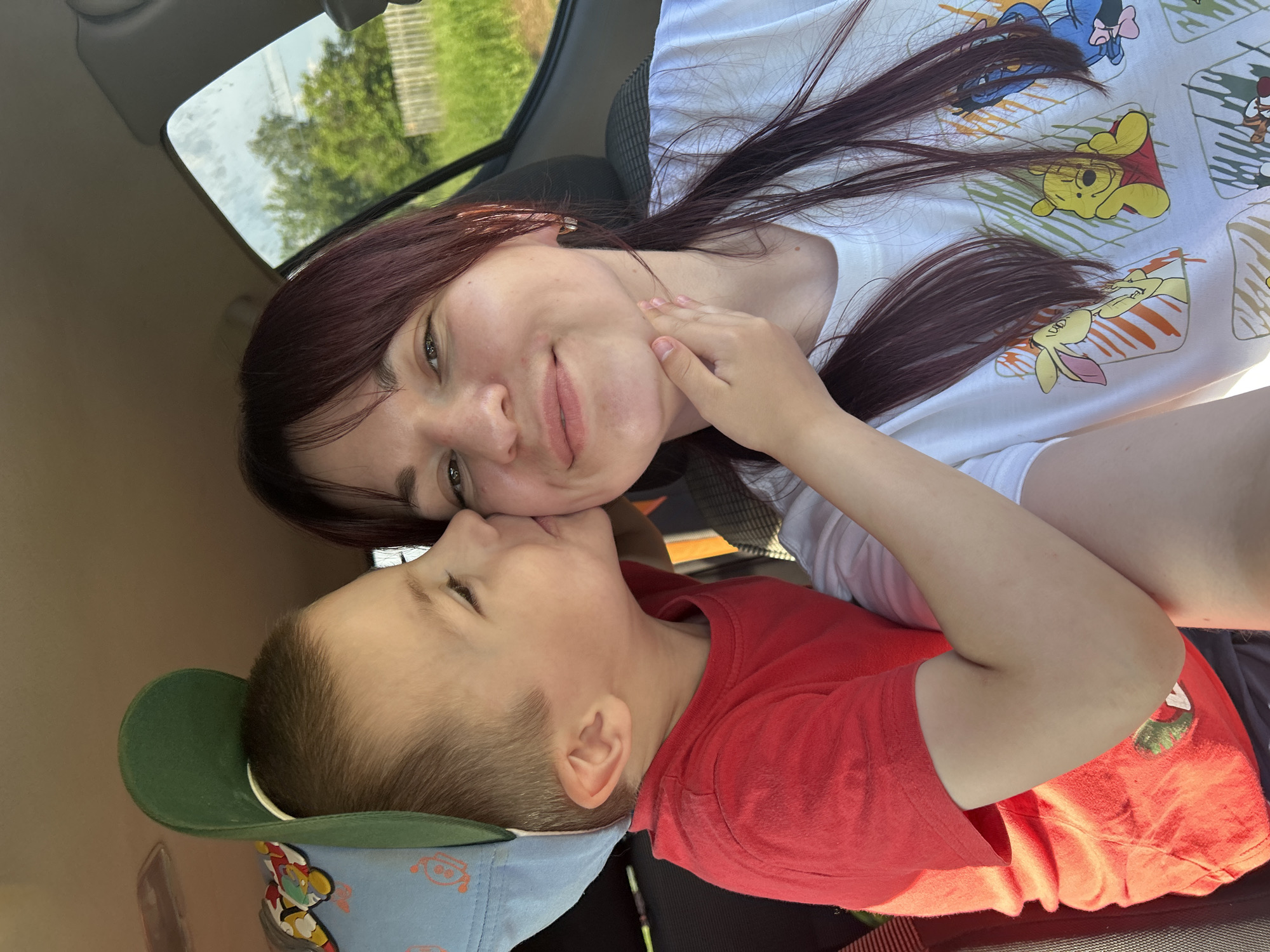 A boy wearing a red t-shirt and a light blue cap with a green brim is kissing a woman on the cheek. The woman has dark hair and is wearing a white t-shirt with a Winnie the Pooh print. They are in the interior of a car. The background shows a sunny day with green trees and a wooden fence, suggesting a rural or suburban setting. The location is the city of Fokino, Russia.