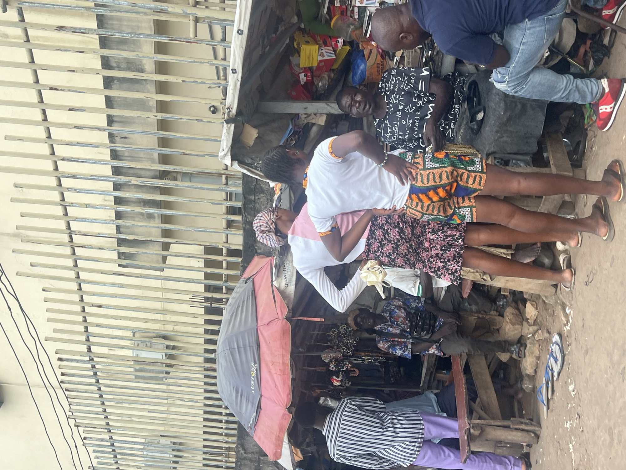 The image depicts a bustling street scene in Nkpor, Nigeria, likely at a market. Several people are gathered around makeshift stalls displaying goods under a large umbrella and tarpaulin. In the foreground, a group of individuals stand and sit, some engaged in conversation or looking at merchandise. One person in a striped shirt and purple pants is standing to the left, facing away from the camera. Next to them, a person in a colorful patterned shirt is seated. Two individuals, one in a pink headscarf and white top with floral skirt, and another in a white top and patterned shorts, are standing and interacting. A seated man in a black t-shirt with scientific formulas is also visible. To the far right, a person in a blue shirt and jeans is seated, looking towards the other individuals. The environment is characterized by wooden stalls, scattered debris on the ground, and a metal fence in the background. The presence of various items for sale, such as jewelry and packaged goods, suggests a commercial setting.