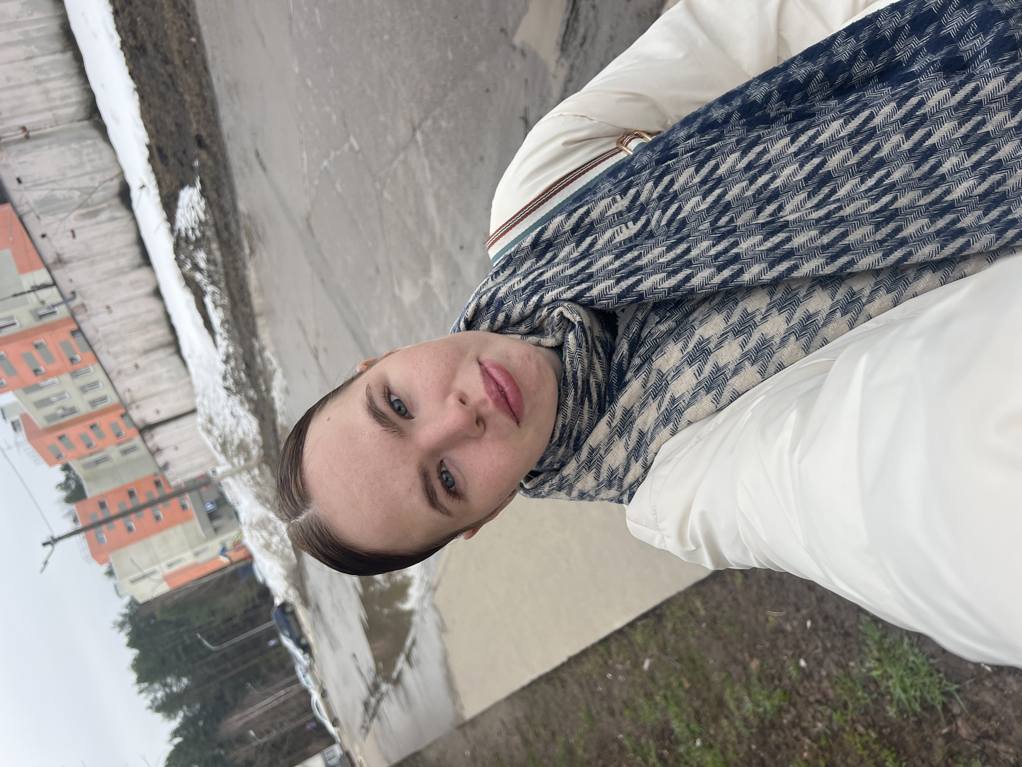 A woman is positioned outdoors, possibly in the city of Chaikovsky, Russia, based on the style of the building in the background. She is wearing a white jacket and a patterned scarf. The environment includes a concrete ground surface, patches of grass, and a body of water reflecting nearby buildings and trees. There is also snow visible near the water. The buildings in the background feature orange and gray facades.