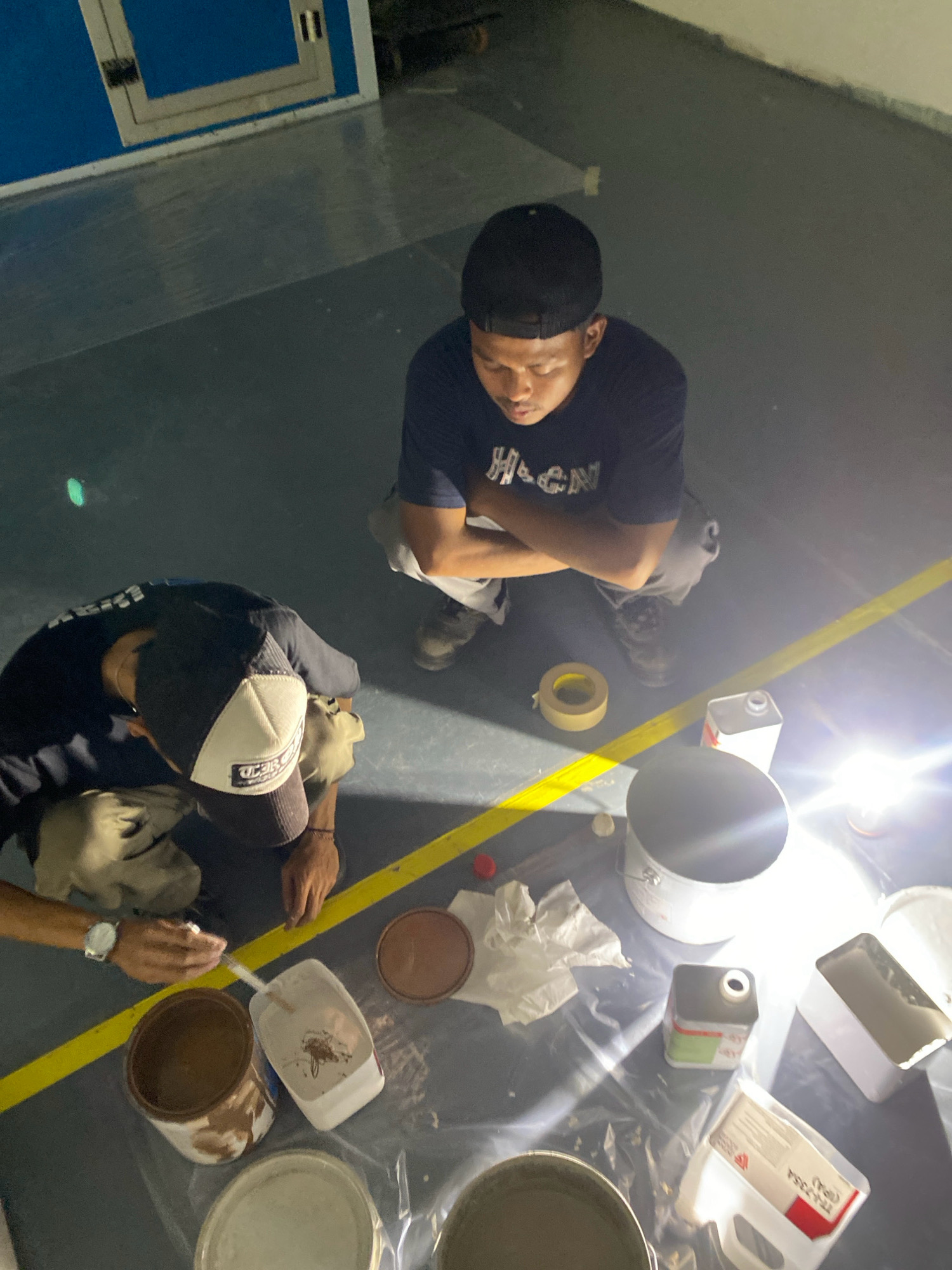 This image depicts two individuals working on what appears to be a floor or ground surface, possibly in an industrial or workshop setting, within the city of Batam, Indonesia. The lighting suggests the scene is indoors, potentially with artificial lighting contributing to the bright flares visible.
One individual, wearing a dark t-shirt with "HIGN" printed on it and a black baseball cap, is crouched with arms crossed. The second individual, wearing a dark t-shirt and a baseball cap with a logo, is also crouched. This individual is wearing gloves and appears to be engaged in a task involving painting or applying a substance, as evidenced by the presence of paint cans, a brush, and a white mixing container. A yellow line is marked on the floor, dividing the space.
Objects present include several open and closed paint cans of various sizes, a roll of tape, crumpled white paper or cloth, and plastic sheeting spread on the floor. One paint can has a brown, viscous substance, while another contains a lighter colored liquid. A white container with a red label and text is also visible. A watch is worn on the left wrist of the second individual. The environment appears to be a flat, dark-colored surface with a blue and white structure in the background. The overall scene suggests a work in progress, likely related to floor maintenance or painting.