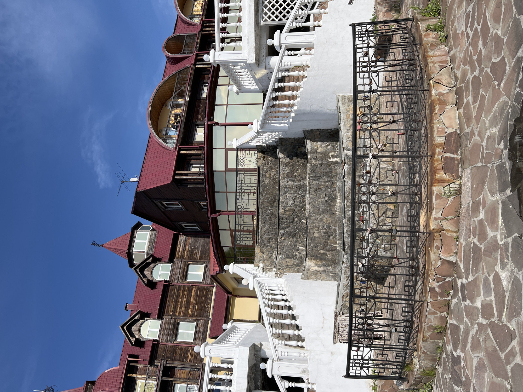A wide-angle, low-angle shot captures a multi-tiered, rustic-style building with red roofs and wooden siding under a clear blue sky. The building is accessed by a grand white staircase with ornate balusters and white globe finials, which is juxtaposed against a cascading stone waterfall feature. Below the waterfall is a black metal railing adorned with numerous love locks. The foreground is paved with irregular, reddish-brown flagstones. The building, known as the "Krestovaya Pad" hotel complex, is situated in Listvyanka, Russia, a settlement on the southwestern edge of Lake Baikal.