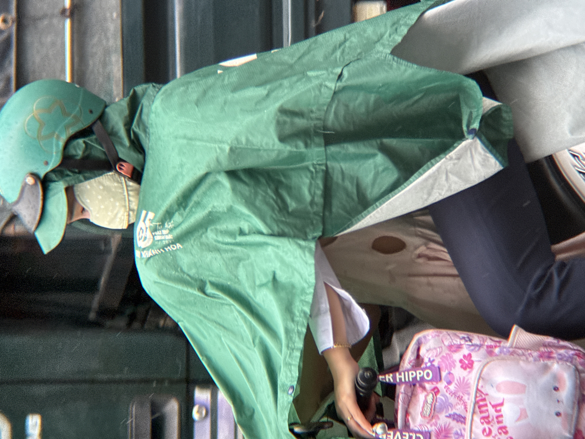 A person on a motorbike in Nha Trang, Vietnam, wears a green helmet and a patterned face mask, covered by a green hooded raincoat. The person is holding the handlebars of the motorbike, with a pink backpack adorned with floral patterns and a rabbit image attached to the front. A white shirt is visible beneath the raincoat. The background consists of dark, indistinct structures, possibly part of a vehicle or building.