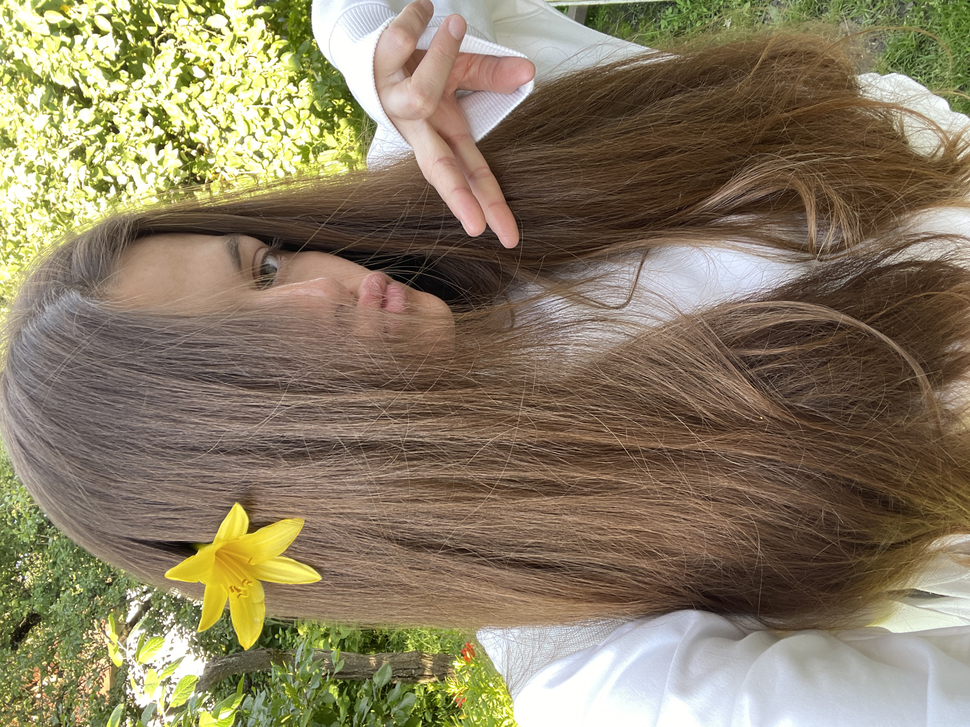 This media file is a close-up, portrait-style photograph featuring a young woman lying down outdoors, with her face partially obscured by her long, brown hair. A single, vibrant yellow flower is tucked into her hair, adding a pop of color to the composition. The woman is wearing a white garment, possibly a hoodie or sweater, with a ribbed cuff visible. Her eyes are open and looking upward, and her lips are slightly parted, suggesting a moment of repose or thought. Her hand is also visible, resting near her head, with her fingers slightly curled. The background consists of lush green foliage and grass, indicating an outdoor setting, likely a garden or park. The lighting suggests it is daytime, with the sun casting gentle shadows. The overall mood is serene and natural. No visible text is present in the image. The location context is Заречный, Russia.