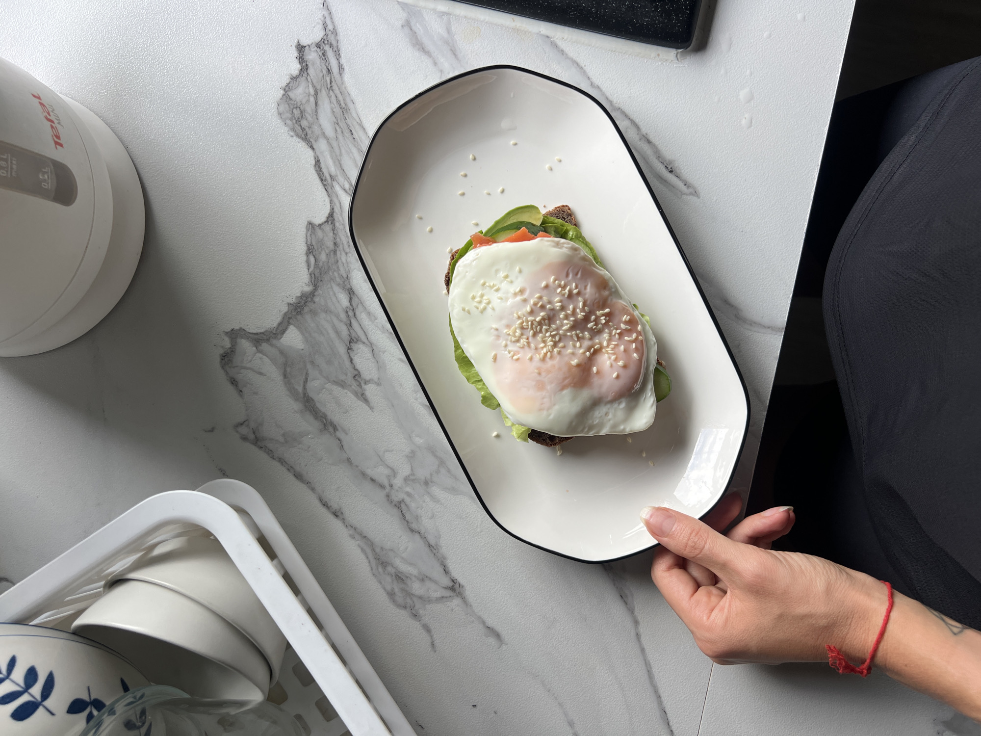 A high-angle shot captures a close-up of a meal presentation, featuring a white, rectangular plate with rounded corners, adorned with a sandwich topped by a fried egg sprinkled with sesame seeds. The sandwich appears to include lettuce, avocado, and tomato. A hand, presumably belonging to a person wearing black clothing and a red bracelet, is seen holding the plate, positioned against a white countertop with a marble pattern. Adjacent to the plate, on the left side, there is a white electric kettle and a dish rack containing white bowls. The location is an indoor setting, likely in a kitchen, situated in the city of Unknown, Unknown.