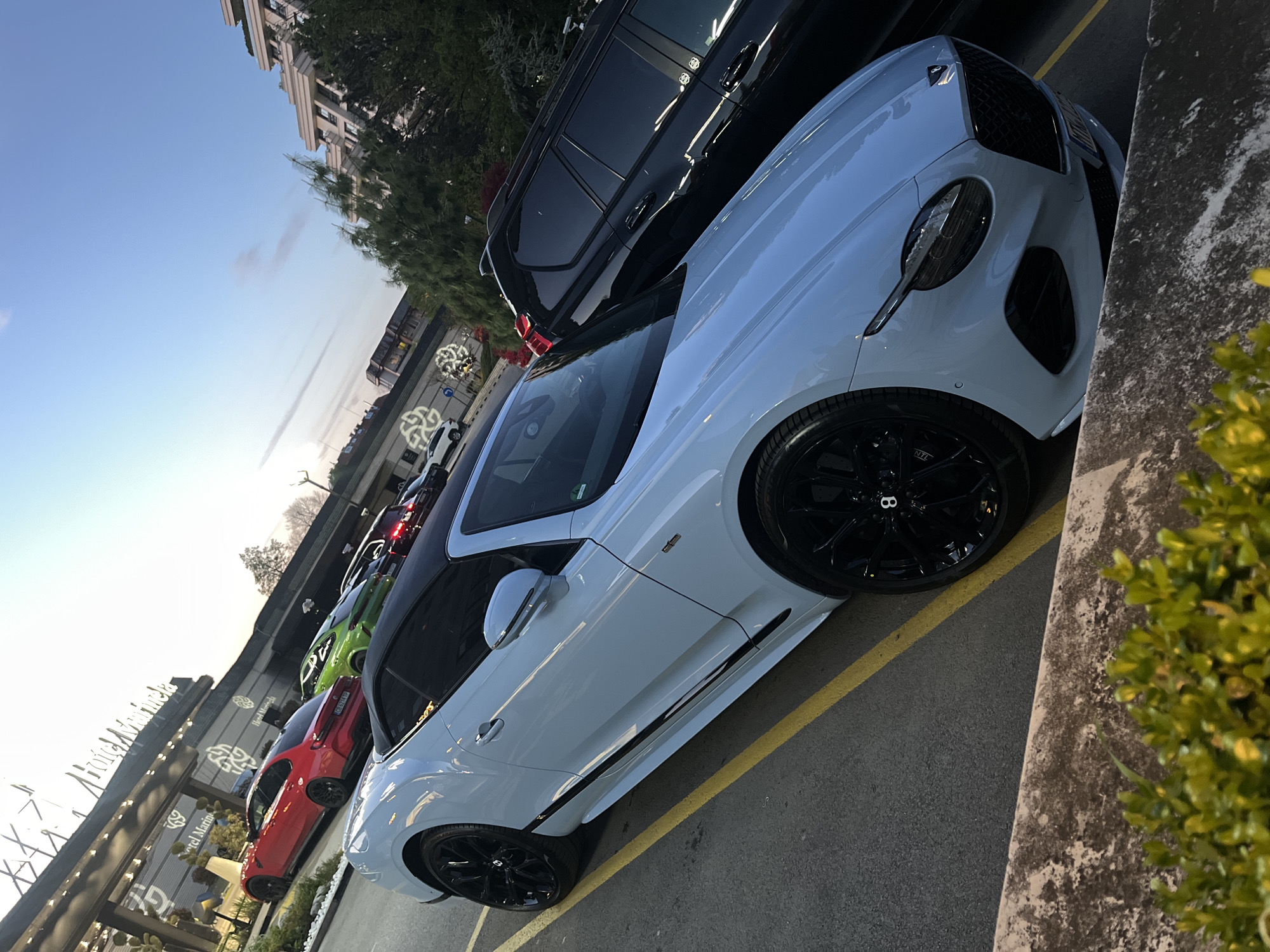 The image shows a row of parked cars, including a light blue Bentley Continental GT with black rims, followed by a black SUV and a red, green, and another red vehicle in the background. The cars are parked along a yellow line next to a concrete barrier with a green bush growing on it. In the background, there is a building with the words "Hotel Marinella" visible. Based on the name of the hotel and the architectural style, the location is likely in Sofia, Bulgaria. The time of day appears to be late afternoon or early evening, as indicated by the sunlight.