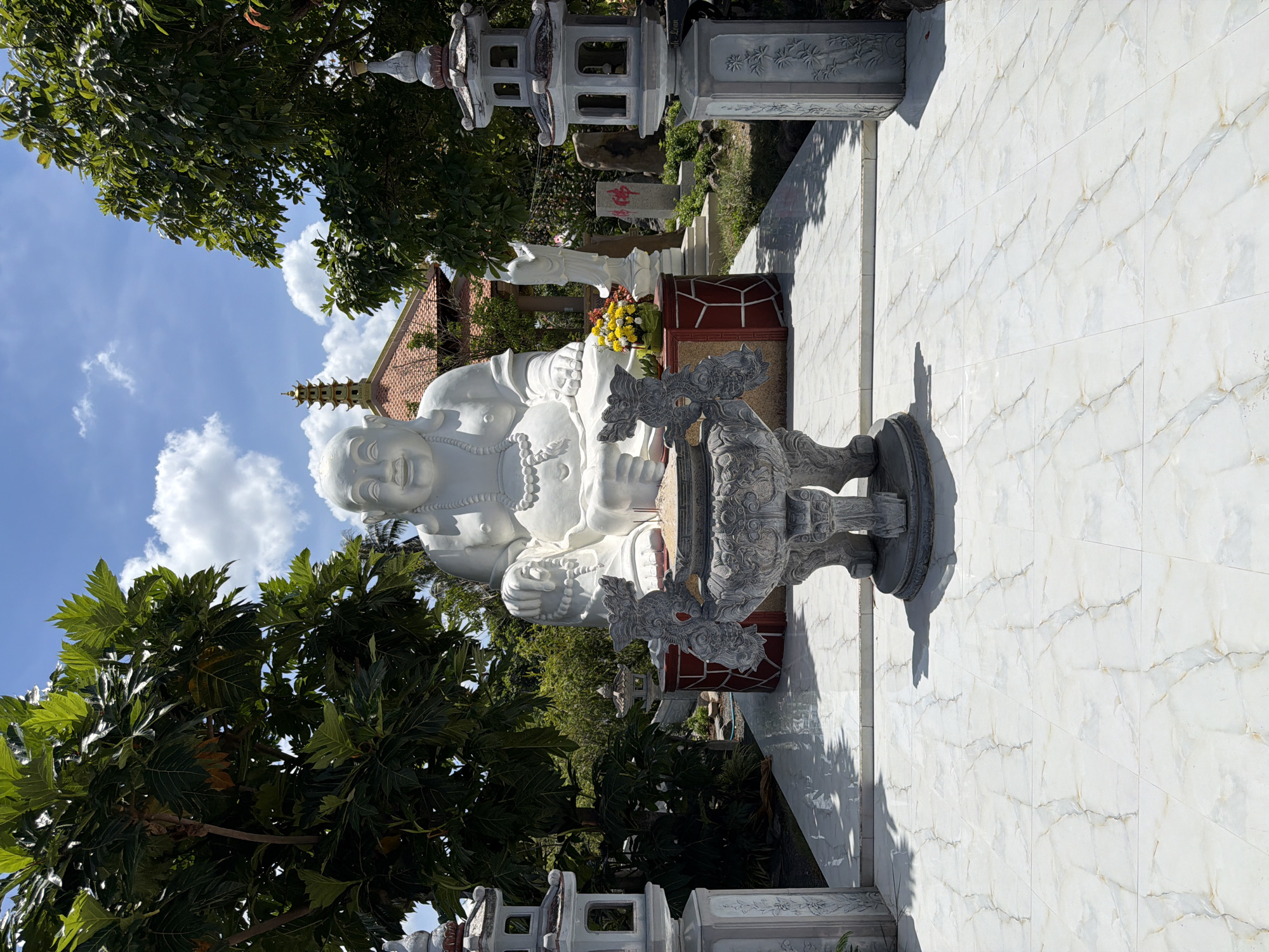The image depicts a serene outdoor scene at a temple or shrine in Tuy An Nam, Vietnam. Dominating the central area is a large, white, seated statue of a jovial Buddha, likely a representation of the Maitreya Buddha or a similar figure. The statue is adorned with a beaded necklace and appears to be made of stone or concrete. In front of the Buddha statue, a stone incense burner rests on a raised platform, with several incense sticks actively burning, emitting a wispy trail of smoke. The platform itself is decorated with dragon-like carvings on the sides of the burner.
Flanking the central Buddha statue are two ornate, multi-tiered white stone lanterns, characteristic of East Asian temple architecture. Behind the Buddha statue, a traditional building with a red tiled roof and intricate detailing is visible, topped by a multi-tiered pagoda. To the right of the Buddha statue, a smaller white statue of a standing figure, possibly a deity or another religious icon, is present. The area is surrounded by lush green trees, with large leaves suggesting a tropical or subtropical climate. The ground in the foreground is paved with large, light-colored marble tiles with a glossy finish, reflecting the sunlight and the surrounding structures. The sky above is a vibrant blue, dotted with fluffy white clouds, indicating a clear day. The overall atmosphere is one of peace and tranquility, characteristic of a religious sanctuary.