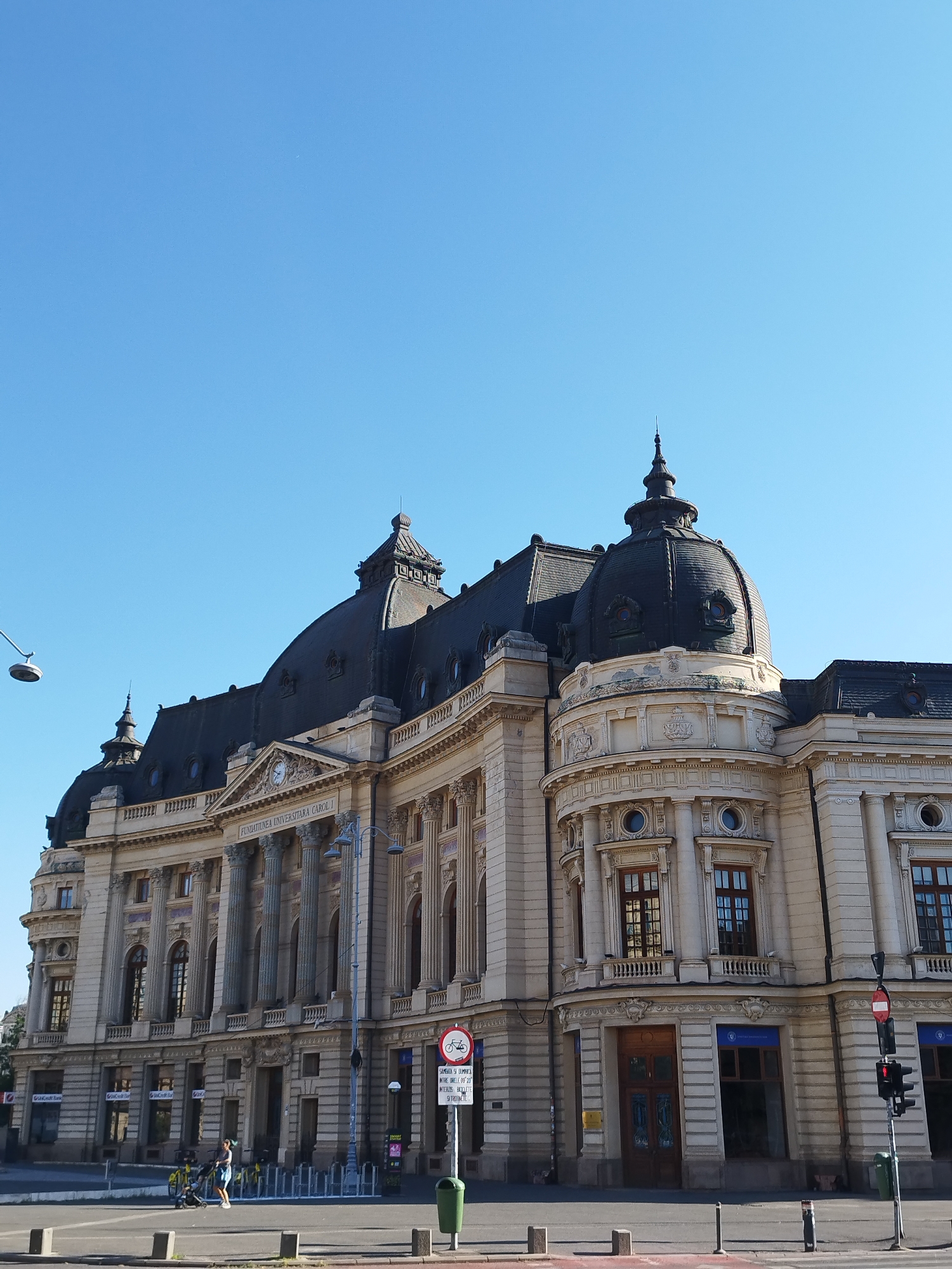 The image captures the monumental facade of the "Fundatia Universitara Carol I" building in Bucharest, Romania, under a clear, bright blue sky. The architectural style is grand, featuring classical elements such as Corinthian columns, ornate carvings, and prominent domes. The building is constructed of light-colored stone, with dark tiled roofs and domed structures, one of which is particularly prominent on the right side of the building.
In the foreground, a pedestrian street is visible, with traffic lights, streetlights, and some yellow rental bicycles parked in a rack. A lone pedestrian, a woman in a blue dress, is pushing a stroller across the street. The street is paved with asphalt, and several concrete bollards are positioned along the edge. A green trash can is situated near the traffic light on the right. The overall impression is one of civic grandeur and a functional urban environment.