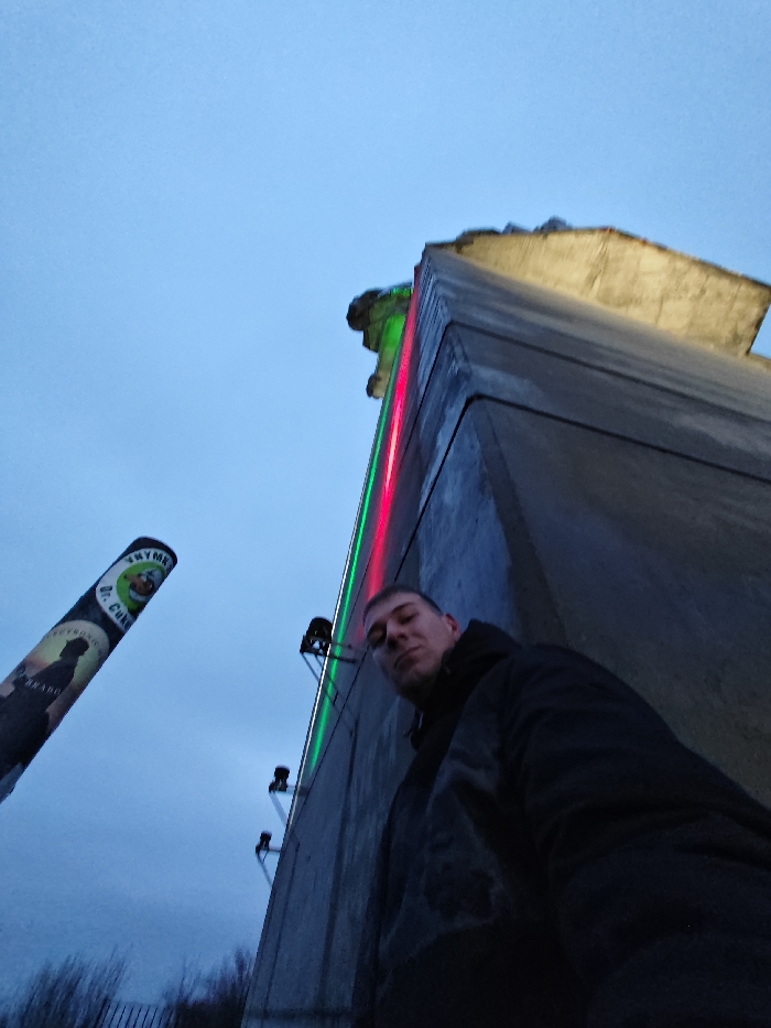 The image depicts a low-angle, outdoor scene at twilight or dusk, featuring one individual in the foreground against the Monument to 1300 Years of Bulgaria in Shumen. The male subject, wearing a dark jacket, occupies the lower-right quadrant, looking directly at the camera with a neutral expression.
The dominant feature on the right is a massive, angular concrete section of the Monument to 1300 Years of Bulgaria. A vertical red light strip is visible above a green light strip along one of its edges. Multiple floodlights are mounted on brackets on the monument's surface, casting upward illumination that highlights a sculpted element near the top of the structure with a yellowish hue.
In the left midground, a cylindrical pole stands vertically, adorned with posters featuring text such as "ВНУЧКИ," "Dr. Coke," and "ANABOL," along with a cartoon character and a silhouette graphic. The sky overhead is a uniform, muted blue-grey, indicative of overcast conditions or twilight. Indistinct dark vegetation and a fence line are visible at the very bottom-left of the frame. The perspective suggests a selfie taken looking upwards towards the monument.