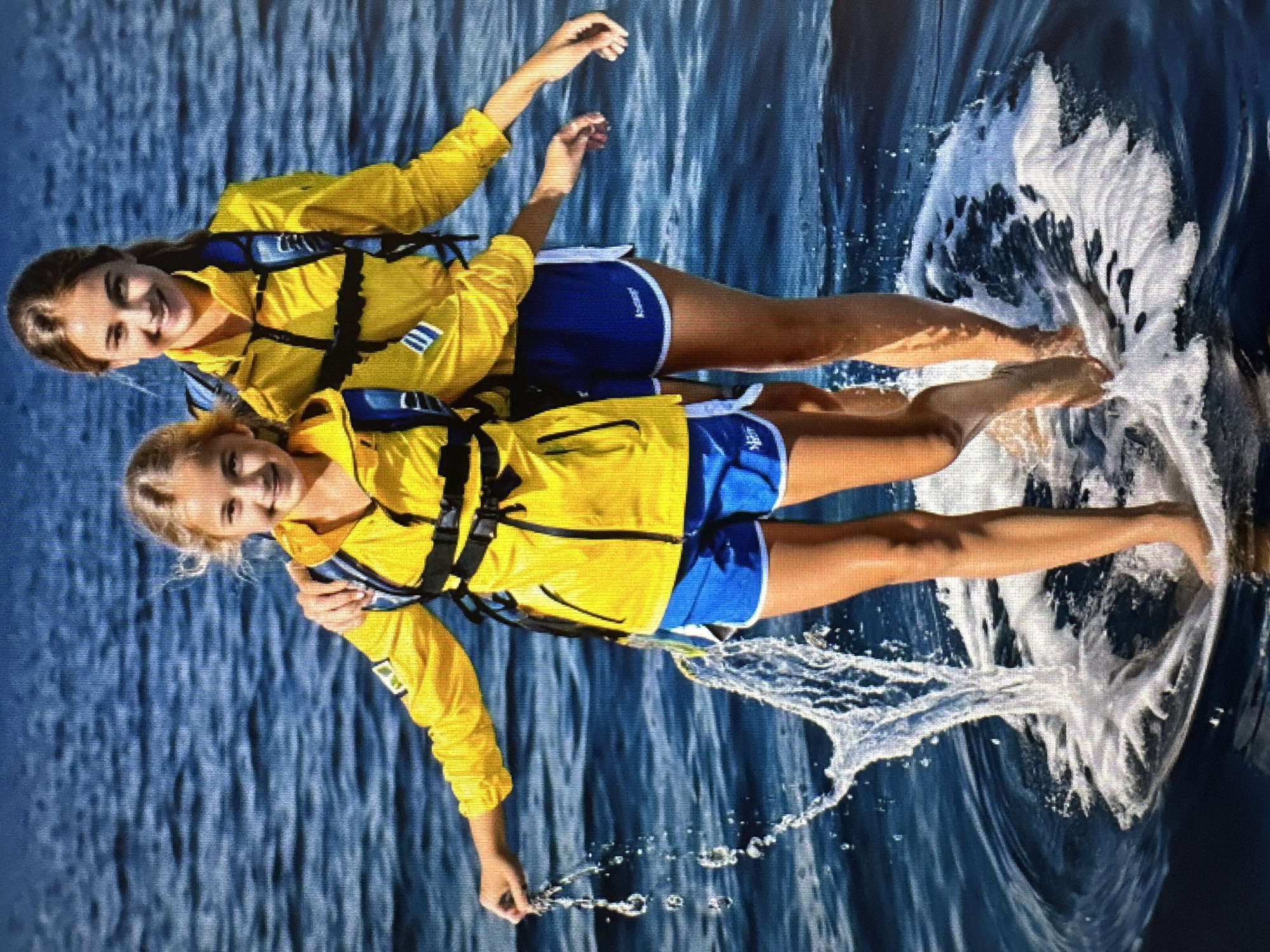 Two young girls, one slightly taller than the other, stand side-by-side on a round, white platform in dark blue water. Both girls are wearing bright yellow jackets over blue shorts. The girl on the left has her left arm around the shoulder of the girl on the right. She is holding a string of water droplets in her left hand, and water is splashing around the platform. The girl on the right has her right arm resting on the left girl's shoulder. Both girls are smiling and looking at the camera. The background is a body of water with gentle ripples.