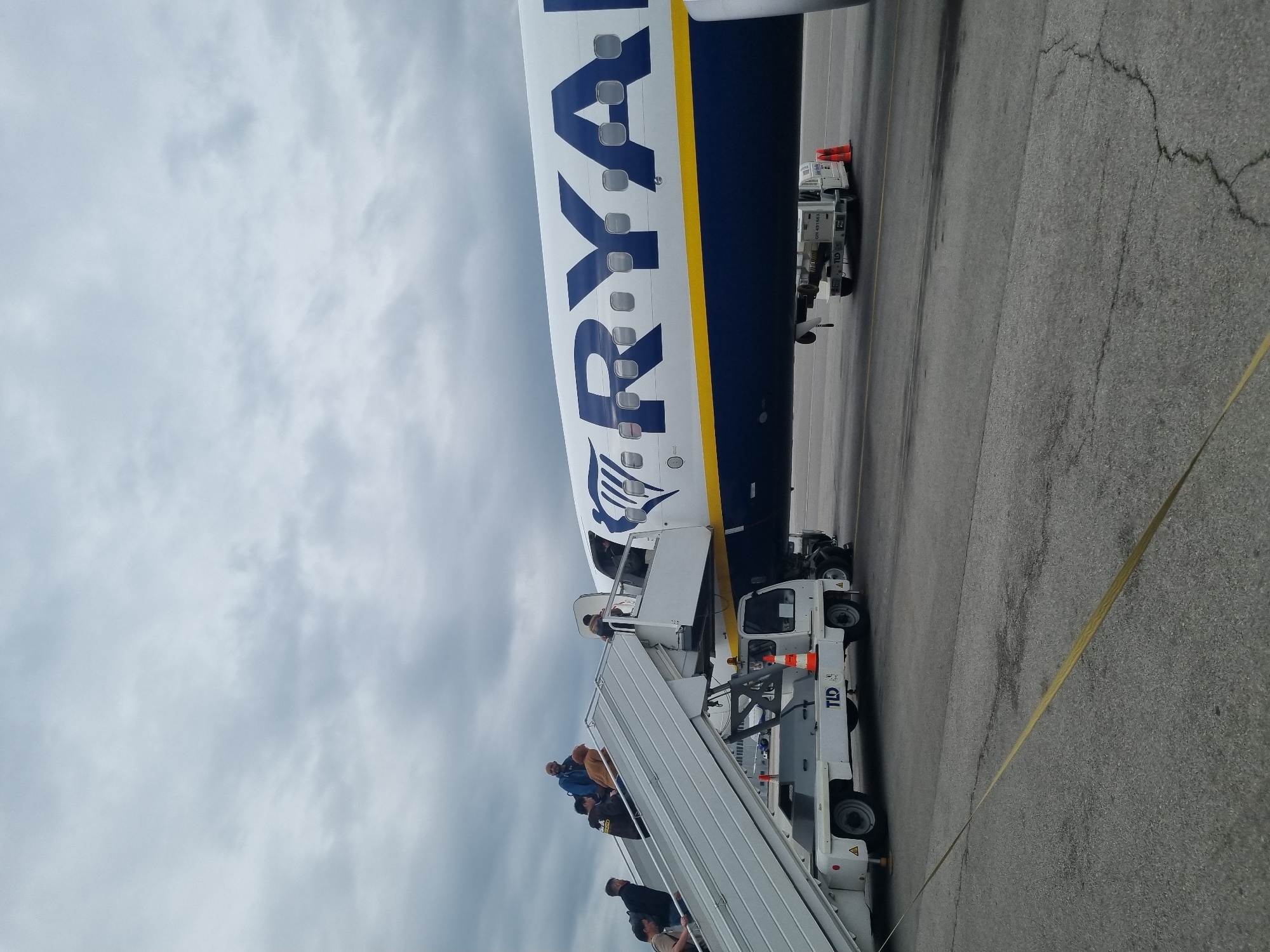 A partially visible Ryanair aircraft is parked on an airport tarmac under a cloudy sky. A set of stairs connected to a white vehicle is positioned at the aircraft's door. Several individuals are descending the stairs. The aircraft's tail and fuselage are blue and white, with the airline's name prominently displayed. The ground surface appears to be asphalt with a yellow line marking. The location is possibly in Sofia, Bulgaria, based on the presence of the Ryanair aircraft.