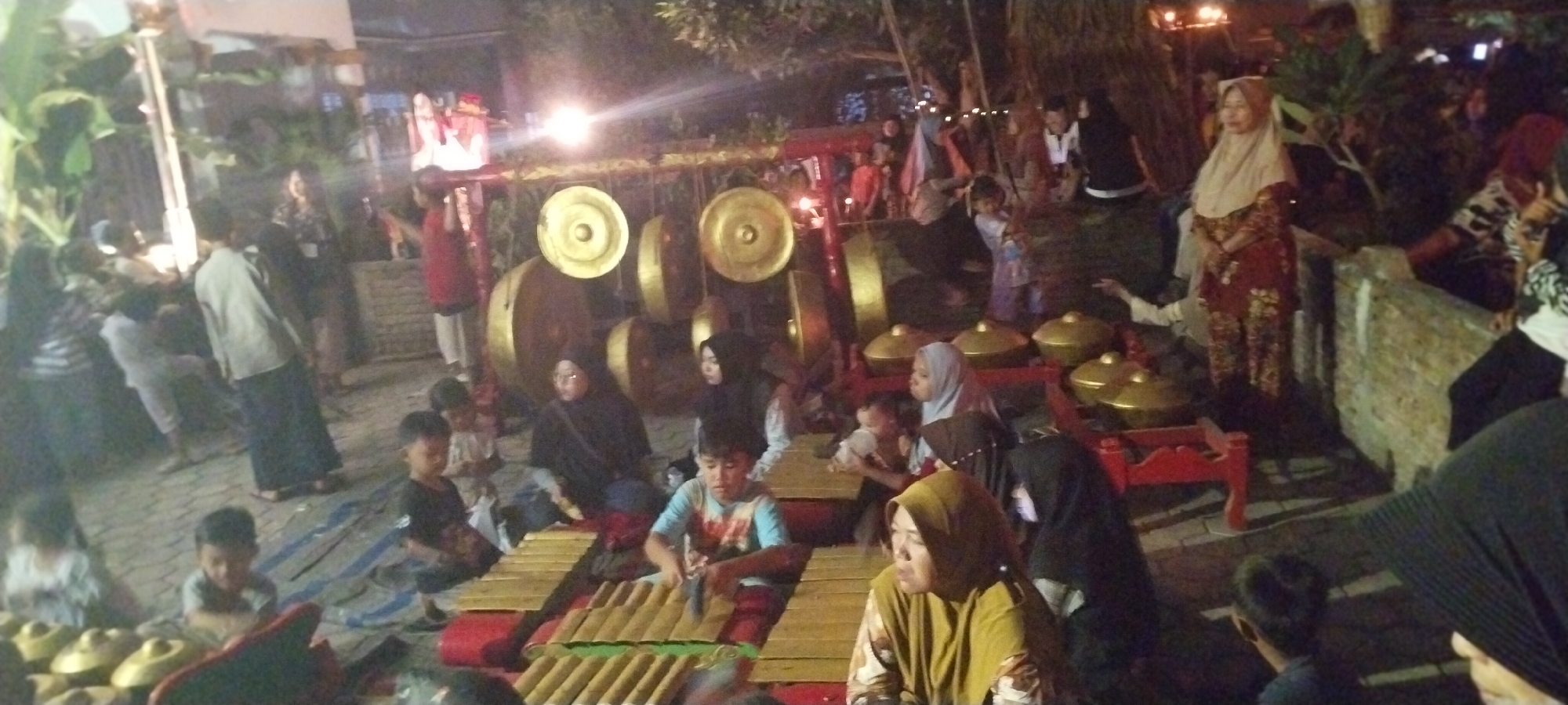 In Keting, Indonesia, at night, a group of people are gathered around a gamelan ensemble. The foreground features a male child playing a xylophone-like instrument, accompanied by other children and adults observing or participating. The background displays a traditional gamelan set with large bronze gongs and smaller instruments, illuminated by artificial lights. People of various ages, some wearing headscarves and traditional clothing, are present. The setting appears to be an outdoor event, possibly a cultural performance or celebration, with trees and foliage visible in the background. The ground is paved with stone and covered with mats where some individuals are seated.