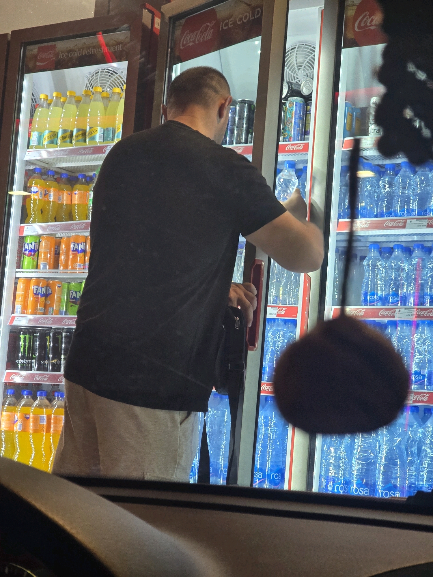 A man with a shaved head and wearing a black t-shirt and beige shorts is standing in front of a refrigerator in a convenience store, located in Podgorica, Montenegro. He is reaching into the refrigerator with his right hand to grab a bottle of water, which is clear and appears to be labeled "rosa". The refrigerator is filled with various beverages, including Fanta, Schweppes, Monster Energy drinks, and many bottles of water. The store has Coca-Cola branding visible on the refrigerator doors and shelves. The scene is viewed from outside the store, through a car window.