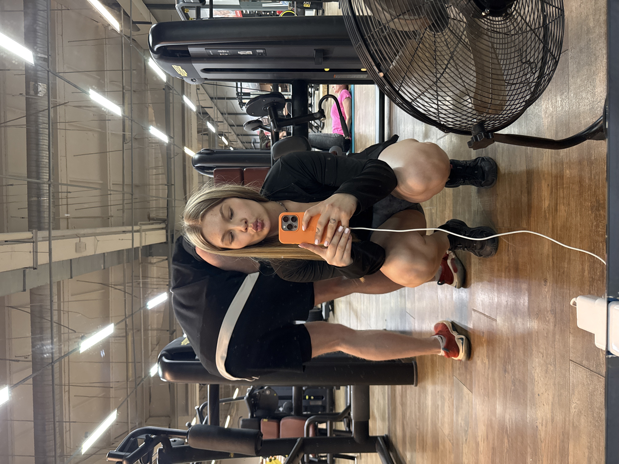 The image, oriented vertically, appears to be a selfie taken with a smartphone in what looks like a gym. A young woman with long blonde hair is making a "kissing" face at the phone camera while squatting, holding the orange-cased smartphone with her right hand. She is wearing a long-sleeved black top.
Behind her, a person wearing a black jacket with white stripes on the sleeves and black shorts is partly visible; only the upper part of the legs is visible, where one leg is wearing a red and white shoe and the other is wearing a black shoe.
The gym setting is evident from the numerous workout machines in the background and the ceiling structure typically found in large indoor spaces. An electrical fan is visible in the upper right corner of the frame. The floor is made of wood.
The location is described to be in the city of Chaikovsky, Russia.