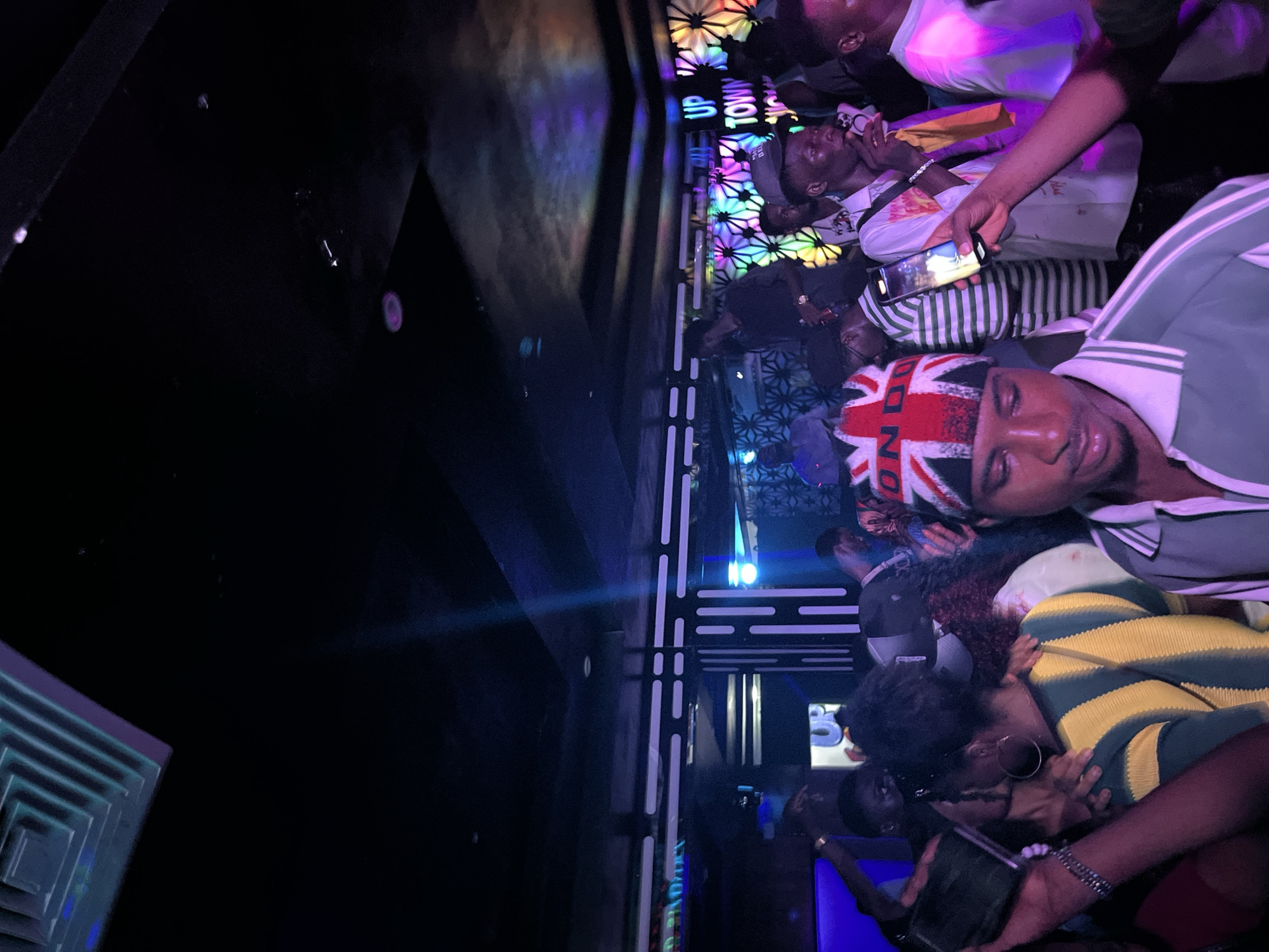 A medium shot captures a group of people inside a dimly lit nightclub. The atmosphere is energetic, with vibrant neon lights casting colorful hues across the scene. The foreground is dominated by a person wearing a beanie with the Union Jack flag pattern. They are looking down, holding a smartphone, with a blurred background of other patrons. In the mid-ground, several individuals are visible, some interacting with each other, while others are looking towards the camera or engaged in their own activities. The backdrop features a wall adorned with a geometric, floral-like pattern, illuminated by colorful lights, and a sign that reads "UP TOWN." The ceiling above is dark, with some visible light fixtures. The overall impression is a lively nightlife scene, with people enjoying themselves in a club environment.