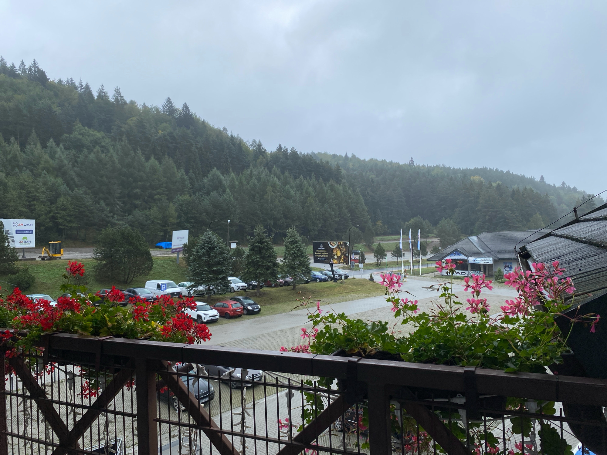 The image captures a view from a balcony overlooking a parking lot and a wooded hillside in Krynica-Zdrój, Poland. In the foreground, a wooden railing supports planters filled with vibrant red and pink geraniums. Beyond the railing, numerous cars are parked on a paved lot. A white van with a blue parking sign is visible. In the middle ground, a few buildings with dark roofs are situated, one of which has "HEAD" signage. Further back, a dense forest covers a sloping hillside under a cloudy sky. Various signs, including a yellow construction vehicle and advertisements, are scattered throughout the scene. The overall atmosphere is overcast and appears to be a cool, possibly damp, day.