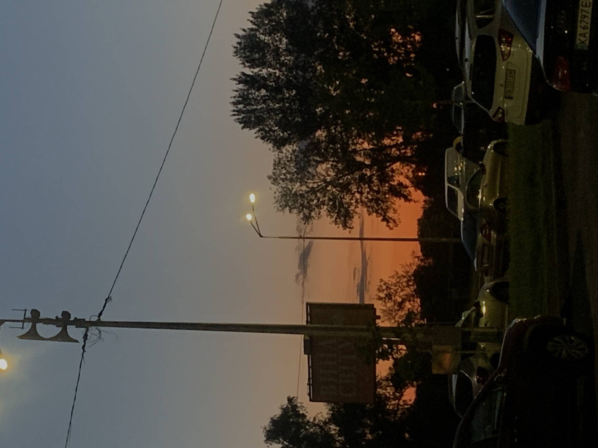 The image depicts a street scene in Kyiv, Ukraine during twilight. A tall pole with what appears to be an emergency siren or speaker system stands prominently in the foreground, attached to a power line. To the right, a street lamp with two illuminated bulbs is visible. In the mid-ground, trees are silhouetted against a colorful sunset sky, transitioning from orange to blue. A billboard with some text, partially obscured by a pole, is situated to the left. The foreground and mid-ground are occupied by several parked cars, some of which are partially illuminated by the street lamp. The overall environment appears to be an urban area with signs of everyday life, indicated by the presence of vehicles and street infrastructure. No people are visible in the frame.