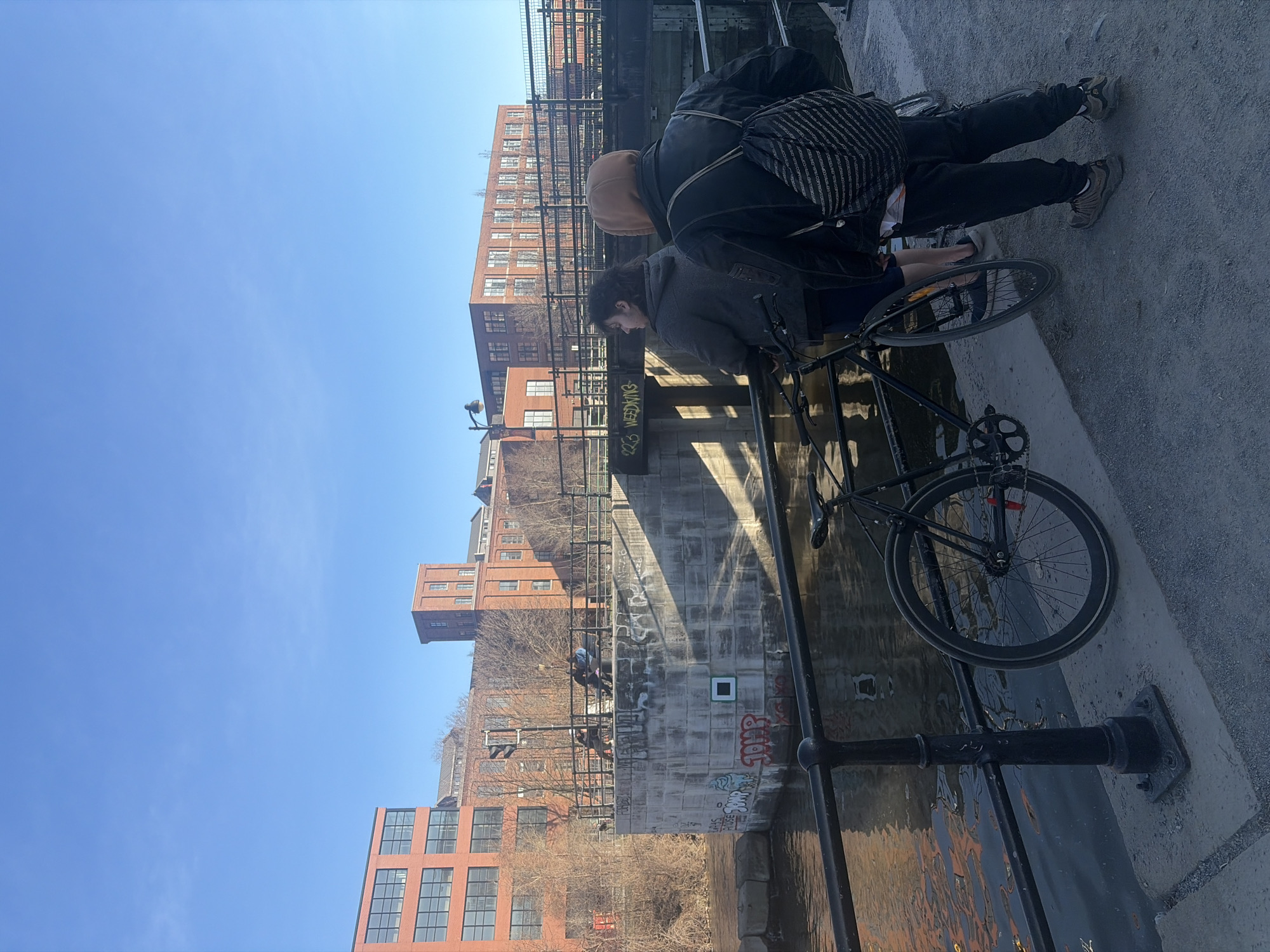 Two individuals are leaning over a bridge railing. A bicycle is resting nearby. The environment features a body of water with buildings in the background. The location is the Lachine Canal in Montreal, Canada.