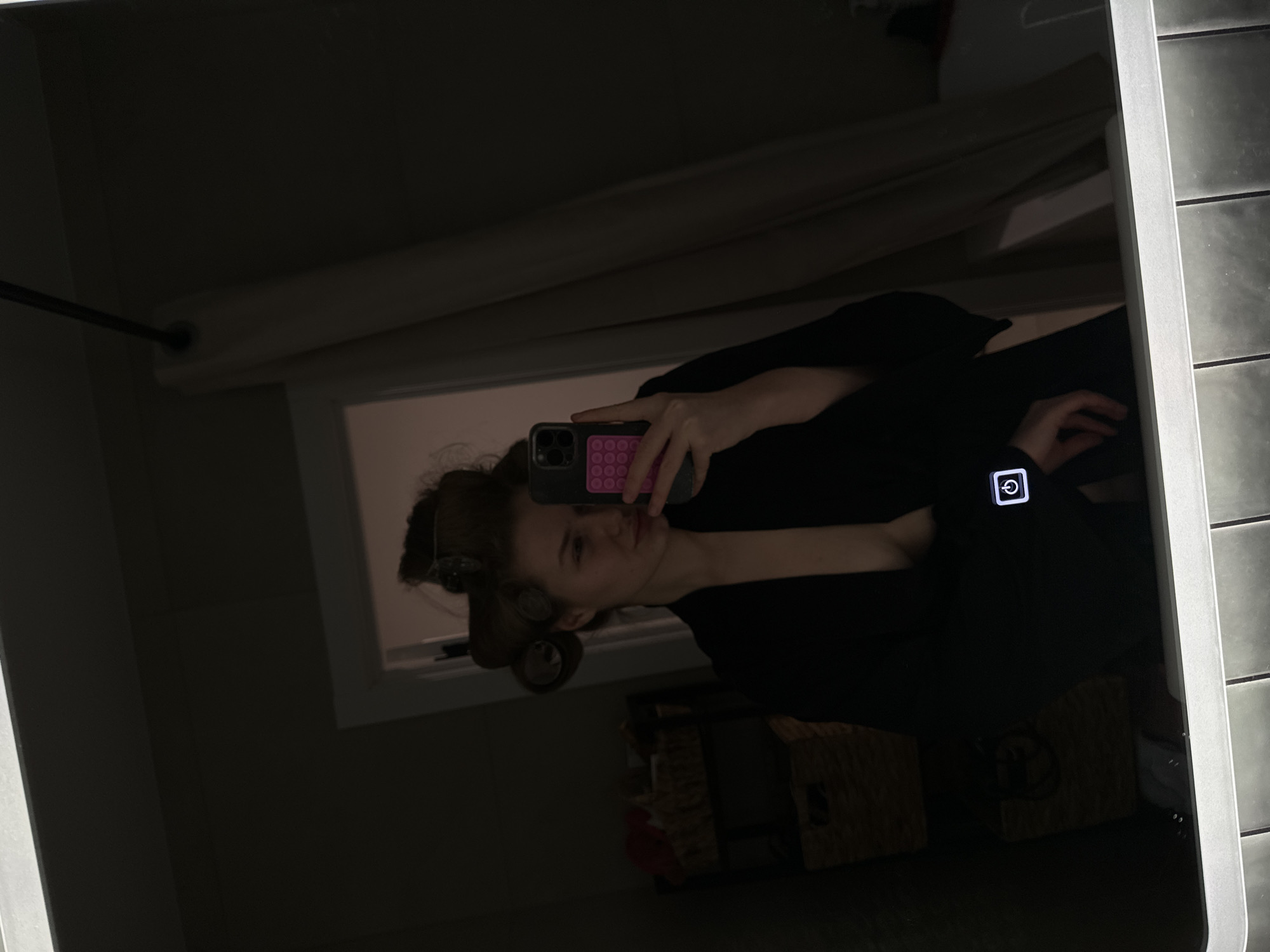 A young woman is taking a mirror selfie. She is wearing a dark robe and has hair rollers in her hair. The reflection shows a bathroom setting with a window. The phone case appears to have a pink pop-it design. The scene shows a modern illuminated mirror and a portion of a dark tiled wall. It is impossible to determine if the location is Kazan, Russia.