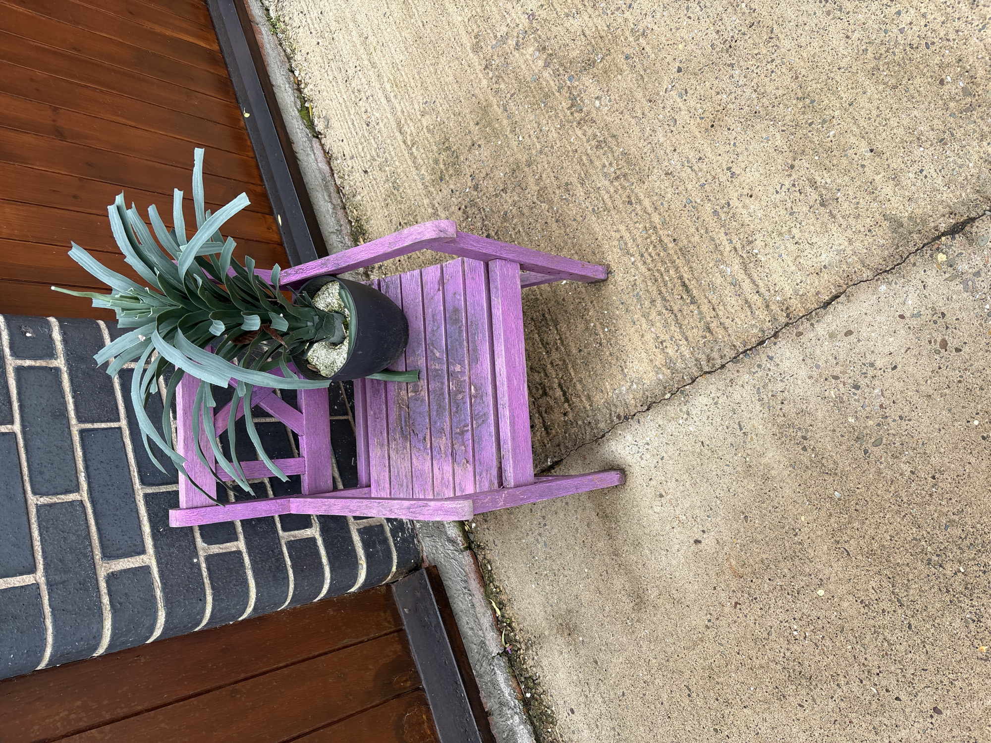 In the city of Pulverbatch, United Kingdom, an outdoor scene is depicted. A weathered, purple-painted wooden chair, with visible paint chipping and discoloration on its seat, stands on a concrete surface. Placed centrally on the chair's seat is a black pot containing an artificial plant with long, flat, greenish-blue leaves. The plant's base within the pot is covered with light-colored pebbles or gravel. To the immediate left and behind the chair is a dark grey brick wall with light-colored mortar, featuring a curved corner. To the right of the wall and behind the chair, a vertical wooden slatted door or gate, painted a dark brown, is partially visible. The concrete ground surface extends from the foreground into the midground, exhibiting a distinct crack that runs diagonally from the lower right toward the middle of the image. The concrete shows signs of wear, including scattered small stones and minor debris. No individuals are present in the scene.