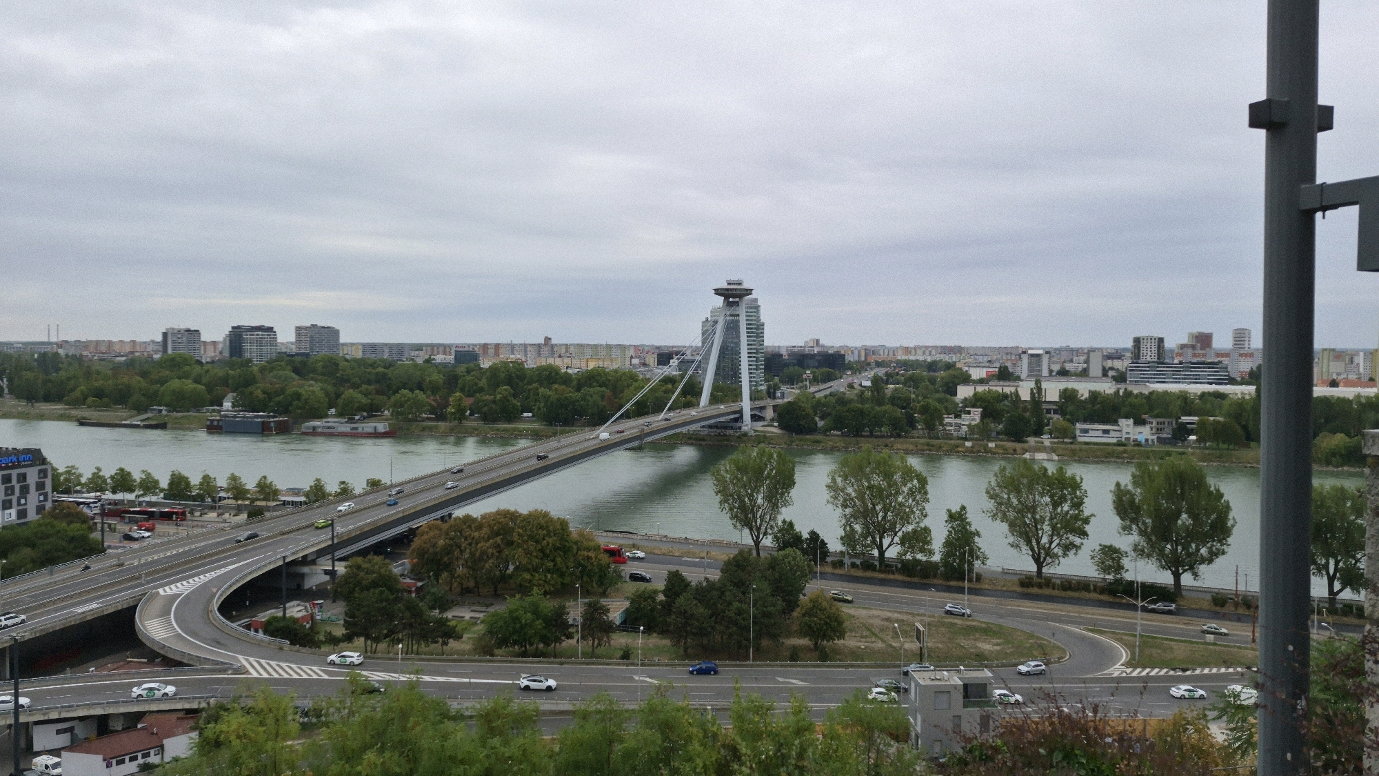 This is a panoramic, slightly elevated, daylight, overcast, outdoor photograph of the Danube river and the city of Bratislava, Slovakia. In the center of the frame is the Most SNP (Bridge of the Slovak National Uprising), a suspension bridge with an iconic UFO-shaped observation deck and restaurant on its pylon. The bridge spans the Danube and connects the historic Old Town to the Petržalka district. Cars are seen driving on the bridge and on the highway ramps leading to it.
The river is a greenish-blue, and on the far bank, a dense line of trees separates the river from a cityscape of multi-story buildings, some appearing to be residential blocks and others more modern office buildings. The sky is a uniform pale gray, indicative of overcast weather. To the right of the frame, a dark gray pole, part of some sort of structure, intrudes into the scene. In the foreground, there are visible trees and a section of road with more cars, some of which appear to be white sedans with green markings, possibly indicating a taxi or delivery service. The overall impression is an urban landscape on a cloudy day.