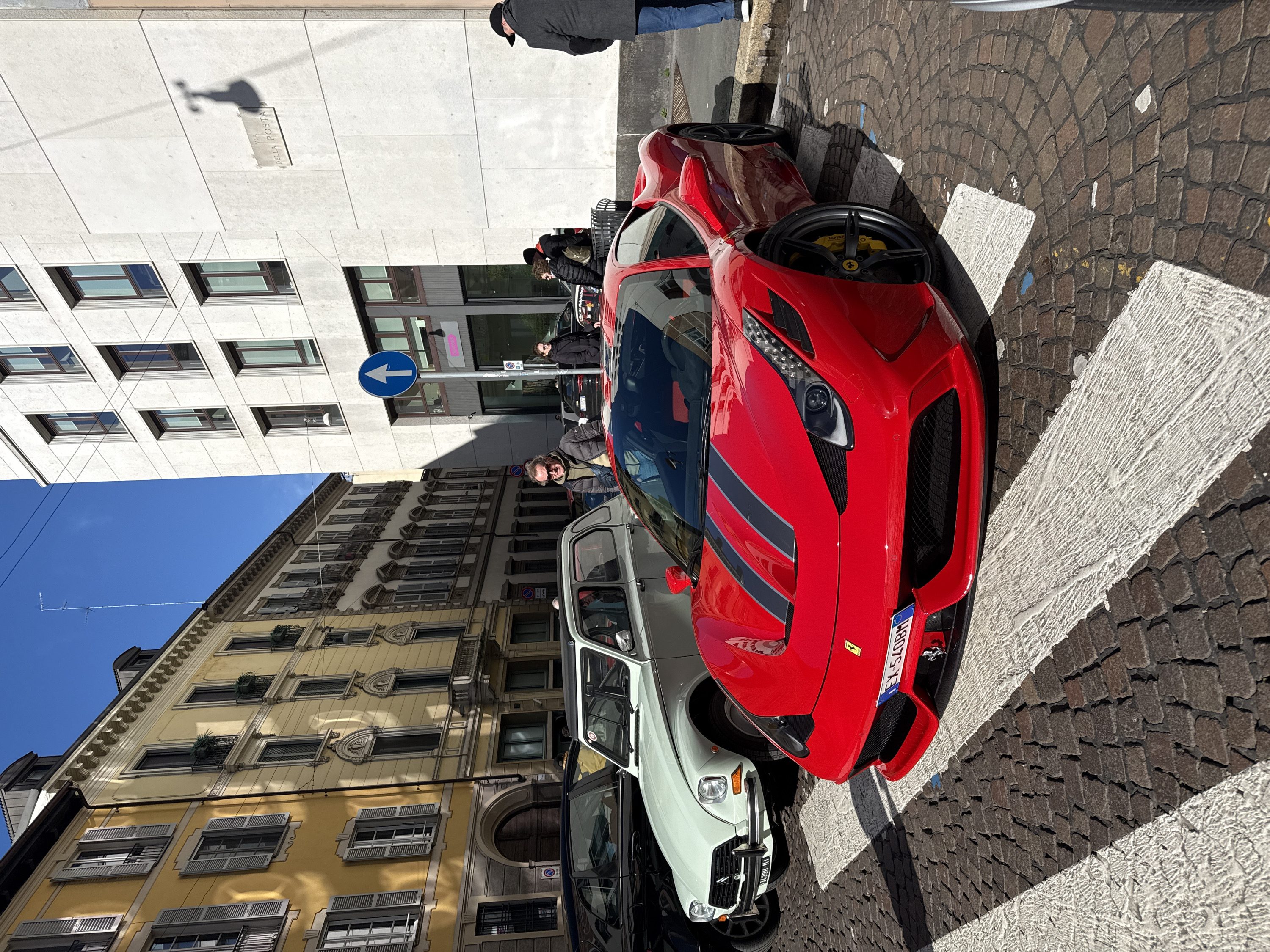 The scene depicts a street in Milan, Italy, specifically Via della Posta, as indicated by a street sign on a modern building. A bright red Ferrari 458 Speciale with black racing stripes outlined in grey on its hood is parked prominently in the foreground, partially on a white pedestrian crossing marked on a cobblestone street. The Ferrari features yellow brake calipers and an Italian license plate "EX-540BH".
Behind the Ferrari, to its left, is a light mint green classic Citroën 2CV with an Italian license plate visible as "34399 HI". A black sedan is parked behind the 2CV.
Several individuals are present:
* A man with sunglasses, a dark puffer jacket, and a green hoodie stands near the Ferrari and Citroën, looking generally towards the camera. He wears a black fanny pack.
* A woman in a dark jacket and glasses stands further back on the right.
* A man in a black cap and dark coat is seen from behind on the far right.
* Other indistinct figures are visible in the background.
The environment includes a mix of architectural styles. On the left, a row of traditional, multi-story buildings with light yellow facades, numerous windows, and light grey shutters are visible. These buildings exhibit classical architectural details like ornate cornices and small balconies. On the right, a contrasting modern, light grey paneled building with dark-framed rectangular windows rises. A blue circular street sign indicating "straight ahead only" is mounted on a pole near this modern building. The sky is clear and bright blue. Strong shadows are cast across the cobblestone street and buildings, indicative of direct sunlight.