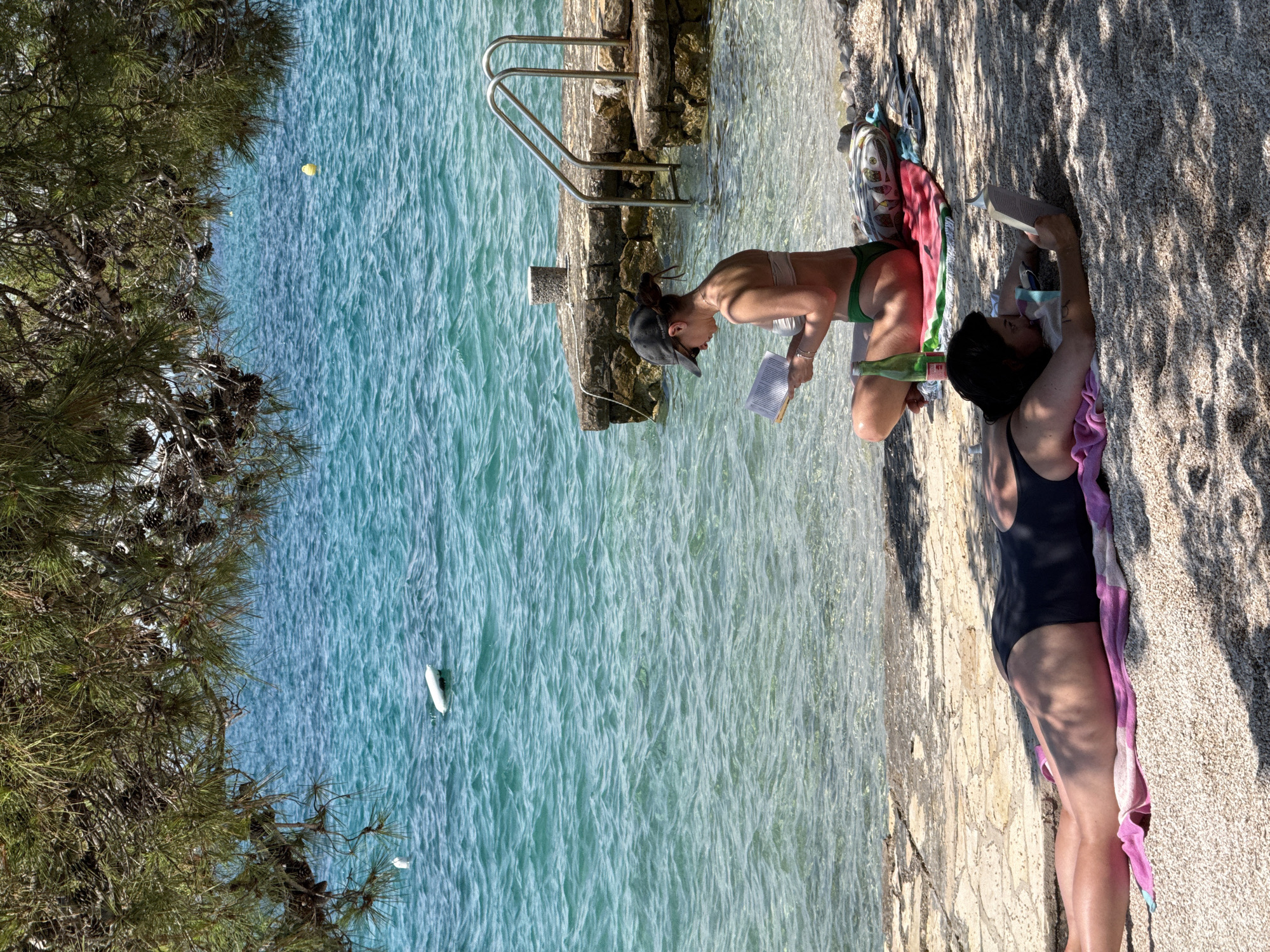The image shows two women at a rocky beach in Grad Šibenik, Croatia. The woman on the left is wearing a green bikini and a baseball cap, and she is sitting in the shallow water, reading a book. She appears to be tanned and has her hair pulled back. To her right, the woman on the left is lying on a towel on the rocky shore, wearing a black swimsuit. She is also tanned and has dark hair. The water is clear and turquoise, and there are some shadows cast by trees on the shore. The time of day appears to be sunny and warm, suitable for a day at the beach. A metal ladder is visible, leading into the water from the rocky shore. There are some pine trees on the left side of the image.