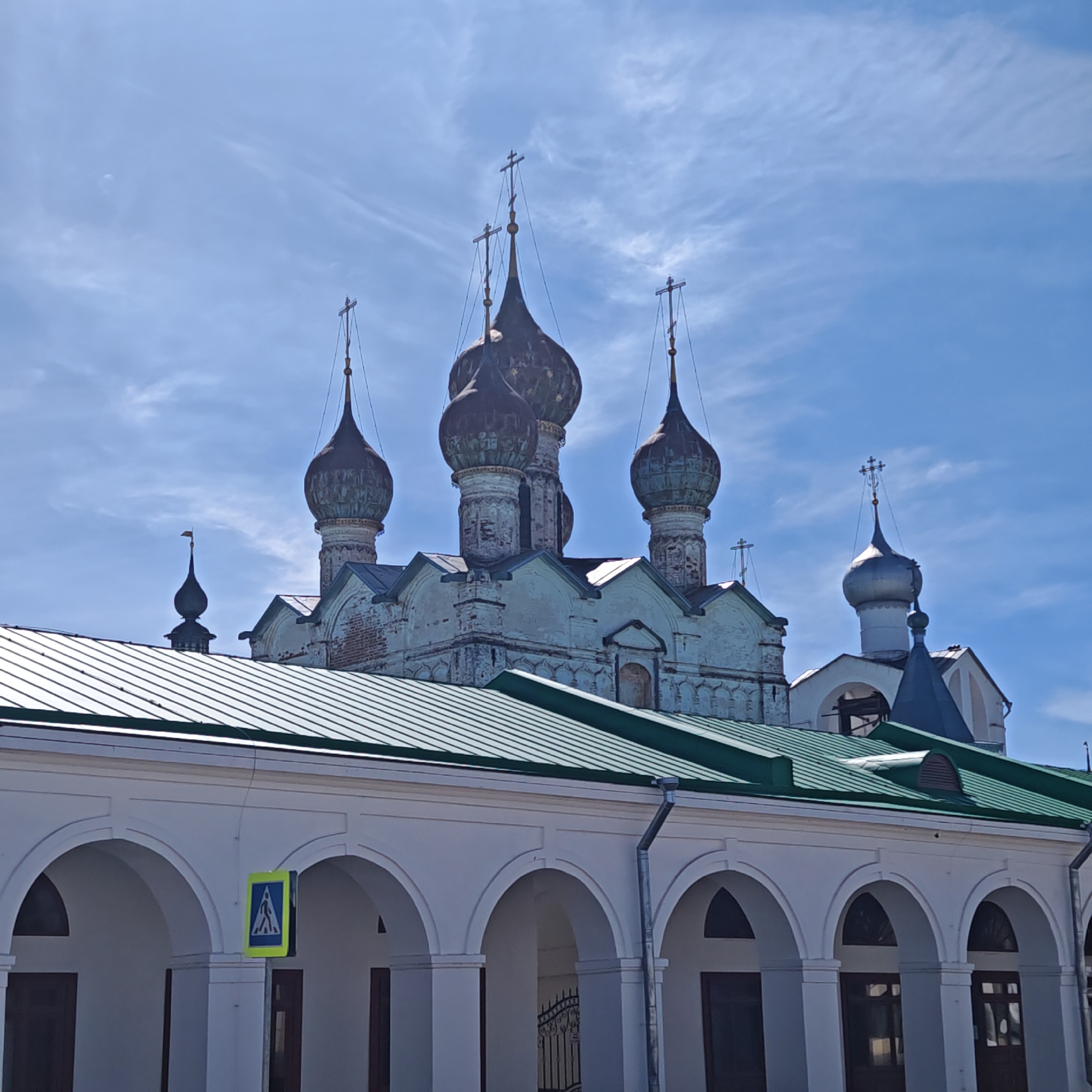 The image depicts a portion of the Rostov Kremlin in Rostov, Russia. Dominating the background is the Church of St. Gregory the Theologian, with its five prominent onion domes, each topped with a cross. The church's exterior appears aged, with visible wear and discoloration. A portion of a separate building with a silver dome and cross is visible to the right. In the foreground is an arcade with white arches and columns, topped with a green metal roof. A pedestrian crossing sign is affixed to one of the columns. The sky is blue with wispy clouds.