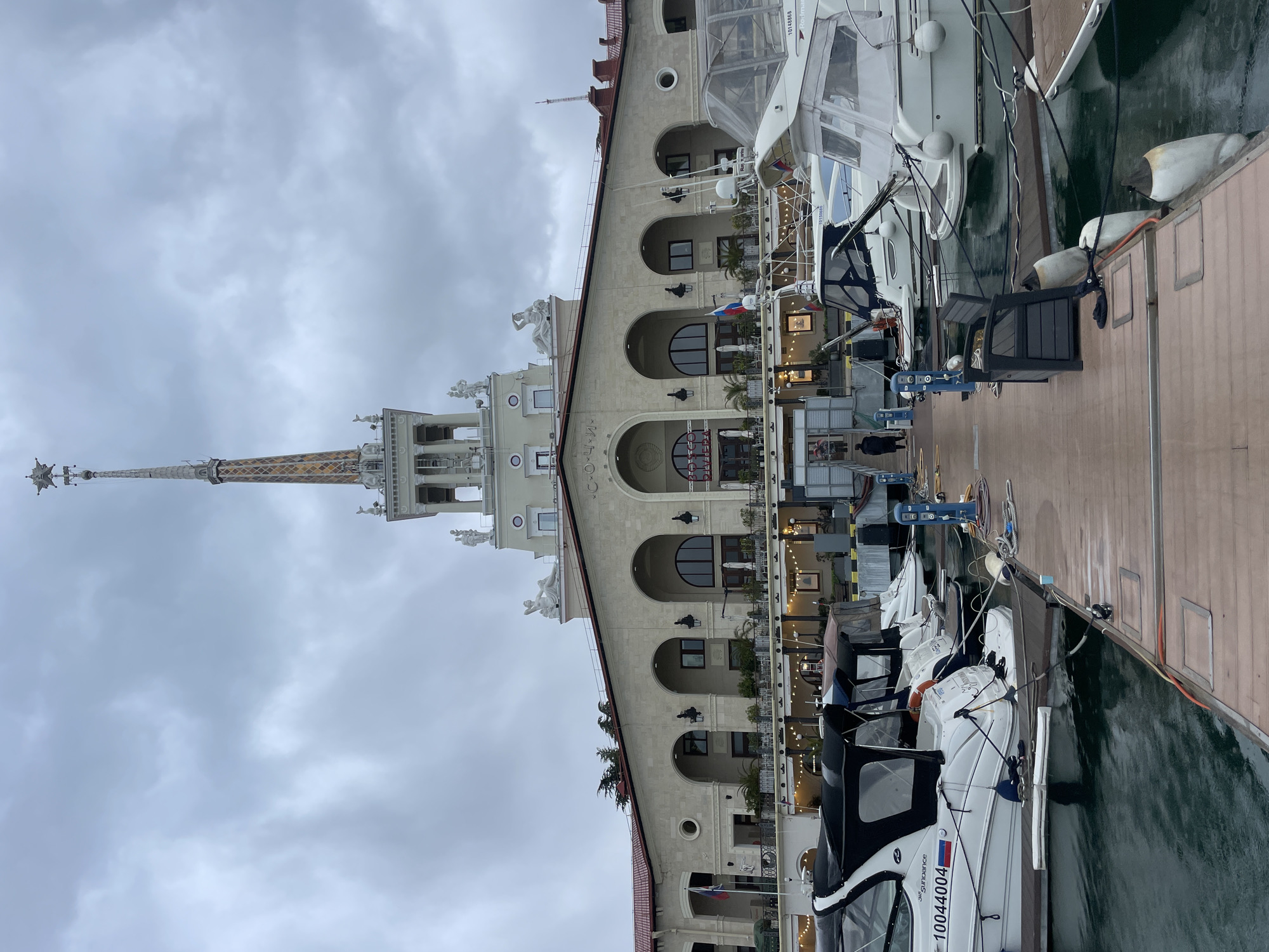 The image shows the seaport of Sochi, Russia, featuring a tall building with a tower and several docked yachts. The building has a light-colored facade with arched windows and decorative elements, including statues. The tower is tall and ornate. The yachts are docked along a wooden pier. The sky is overcast. The scene appears to be a typical view of the Sochi seaport, likely taken on a cloudy day. One of the boats has the number 10044004. There is a Russian flag visible attached to a building.