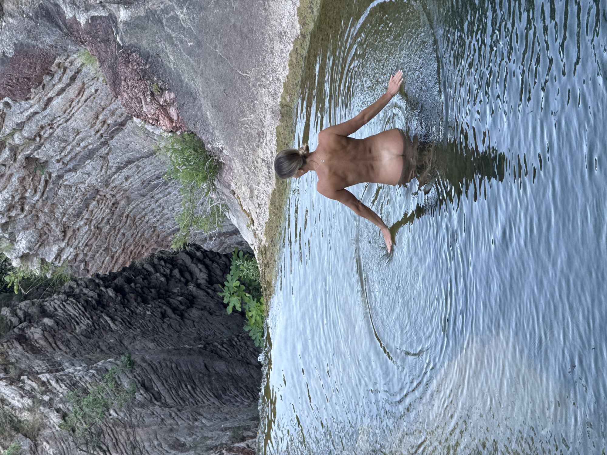 A naked woman stands in shallow, calm water, facing away from the camera. She is positioned in the lower half of the frame, with her hands resting on the water's surface, creating ripples. The water reflects the sky and the surrounding environment. Behind her, a steep, rocky cliff face rises, with patches of green vegetation growing from crevices. The cliff has distinct horizontal striations, indicating layers of rock. The overall setting appears to be a natural, outdoor environment, possibly a secluded swimming spot or a natural pool formed by the landscape. The city of Rafailovići, Montenegro is not depicted in this image.