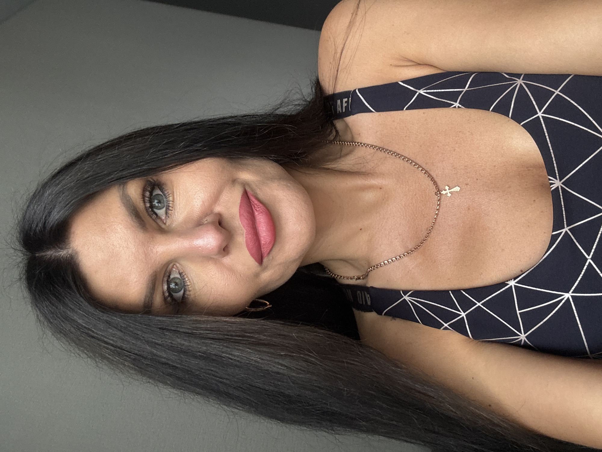 A medium close-up selfie of a woman with long, dark hair and blue eyes. She is wearing a dark blue athletic top with a white geometric pattern. She has on a gold chain necklace with a small cross pendant and hoop earrings. Her lips are painted a bright pink. The background is a plain gray wall. This image was taken in Chaikovsky, Russia.