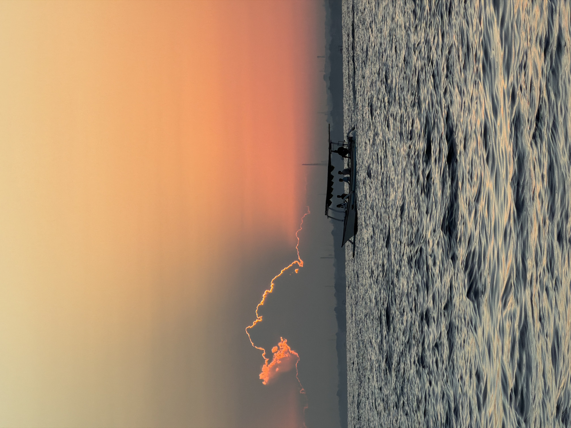 A wide shot captures a boat with five passengers sailing on dark blue, rippling water. The boat, white with a brown canopy, is positioned in the lower-middle section of the frame. Silhouetted against a vibrant orange and yellow sky, the passengers appear to be enjoying a sunset cruise. In the distance, a hazy coastline is visible, with the faint outlines of industrial structures and trees dotting the horizon. A single, large cloud formation on the left side of the sky is brightly illuminated by the setting sun, creating a dramatic contrast with the darker sky behind it. The overall atmosphere is serene and peaceful, typical of a tropical sunset in Indonesia.